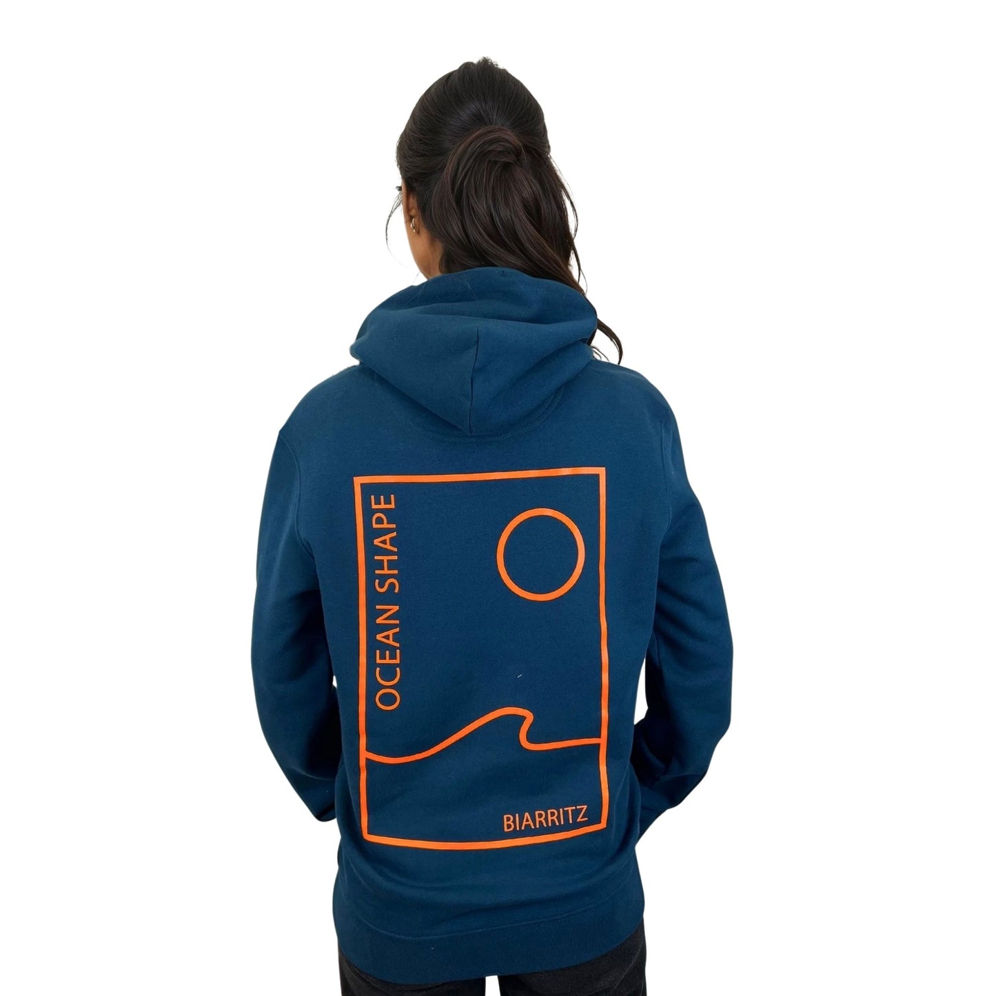 Sweat unisexe à capuche peacock blue avec petit encadré vague / soleil minimaliste + écriture "free as ocean" orange fluo sur la poitrine, vu de face sur mannequin femme
