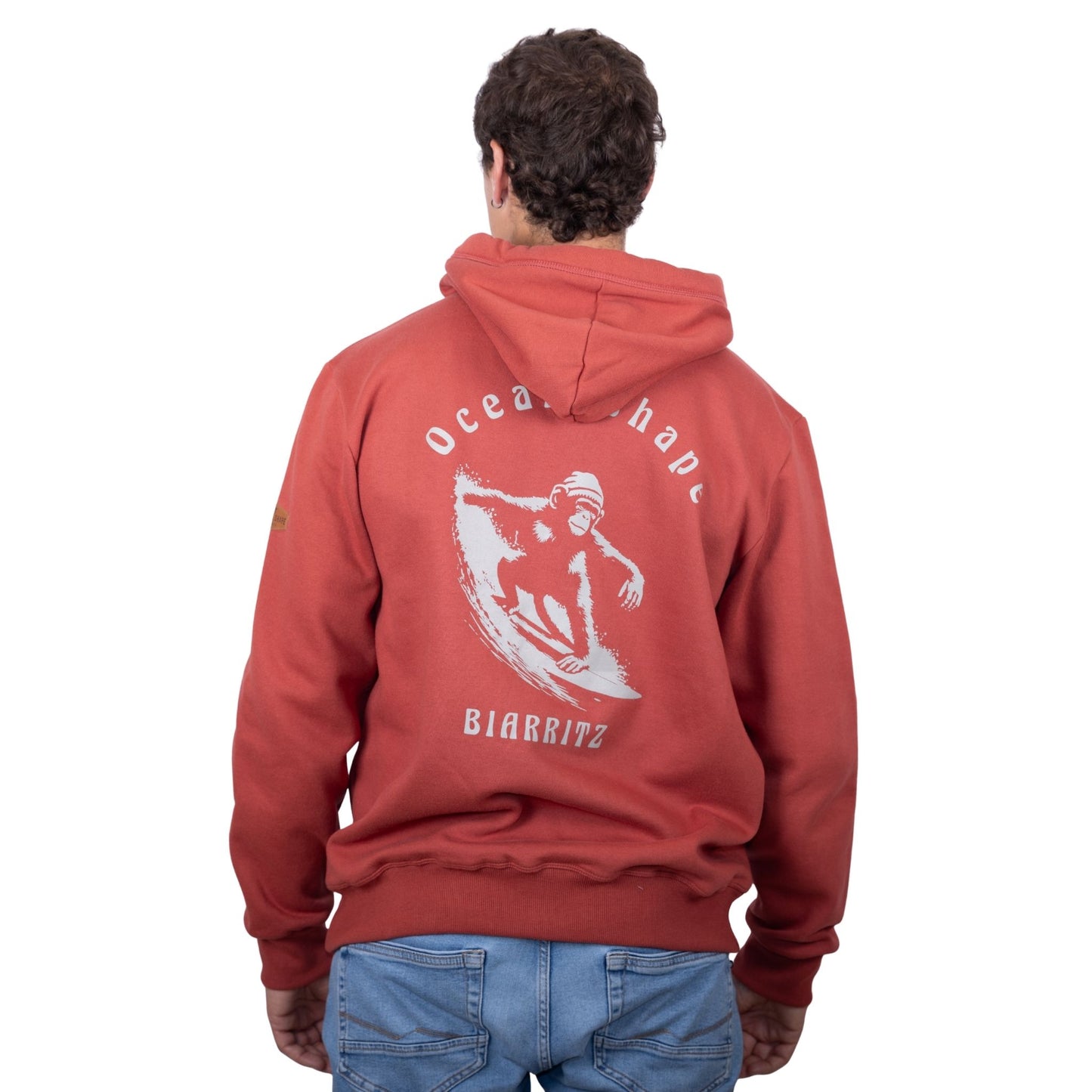 Sweat unisexe à capuche terracotta avec grand dessin de singe qui surf + écriture "Ocean Shape Biarritz" beige dans le dos, vu de dos sur mannequin homme