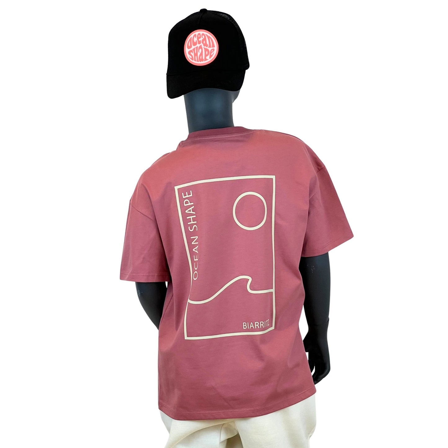 t shirt à manches courtes enfant unisexe couleur anthique rose, en coton biologique, sur un mannequin enfant de face.
Dos : grand dessin minimaliste d'une vague et d'un soleil avec écrit "ocean shape biarritz"
Le mannequin porte une casquette suédé noir ocean shape