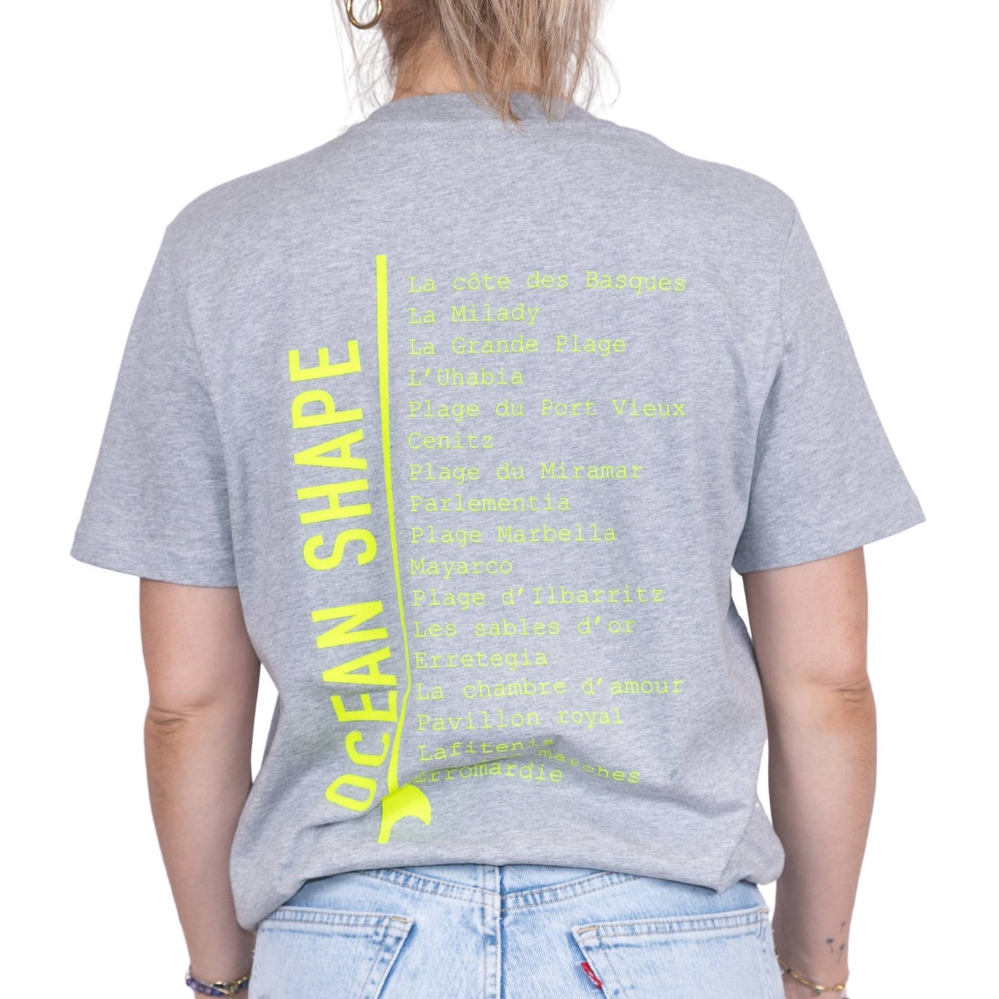Tee-shirt unisexe gris clair en coton biologique avec longue liste de plages et grande écriture "ocean shape" jaune fluo dans le milieu du dos, vu de dos sur mannequin femme