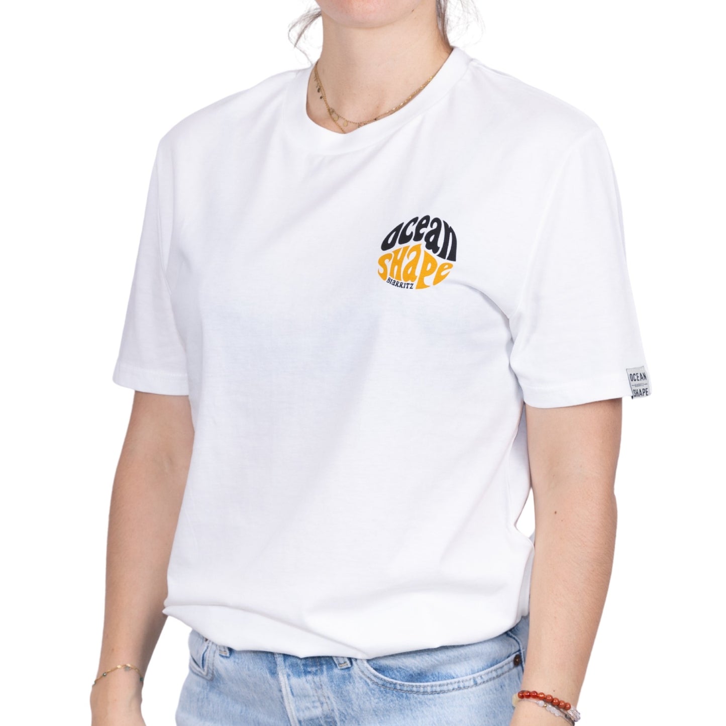 Tee-shirt unisexe blanc en coton biologique avec petit logo ocean shape Biarritz bubble noir et moutarde sur la poitrine, vu de face sur mannequin femme