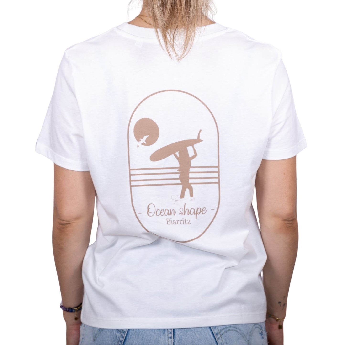 Tee-shirt femme en coton biologique blanc avec gros dessin de surfeuse qui marche de dos et écriture "ocean shape Biarritz" beige dans le dos , vu de dos sur mannequin femme