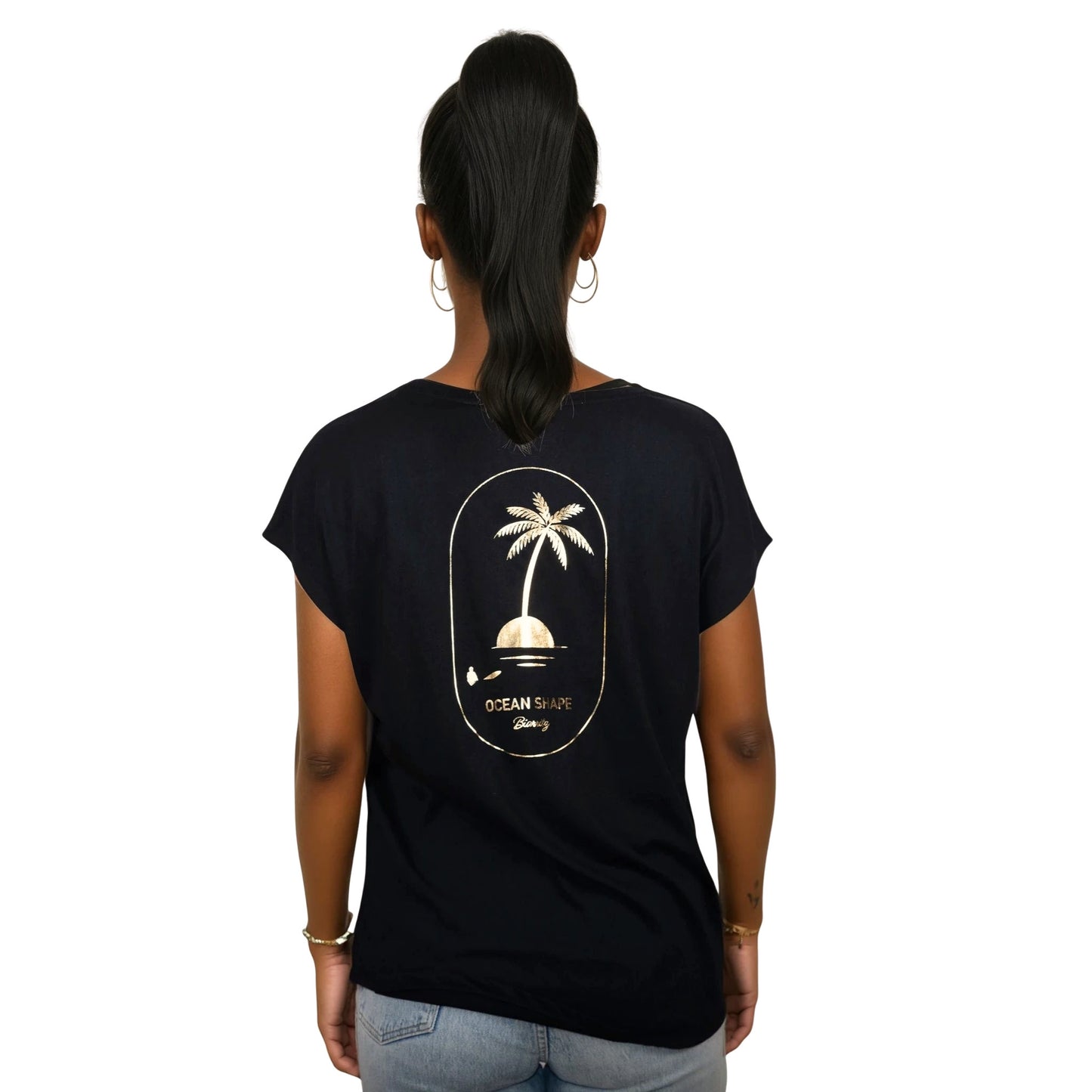 Tee-shirt femme en coton biologique noir avec gros dessin palmier sur île et écriture « ocean shape Biarritz » couleur doré dans le dos, vu de dos sur mannequin femme