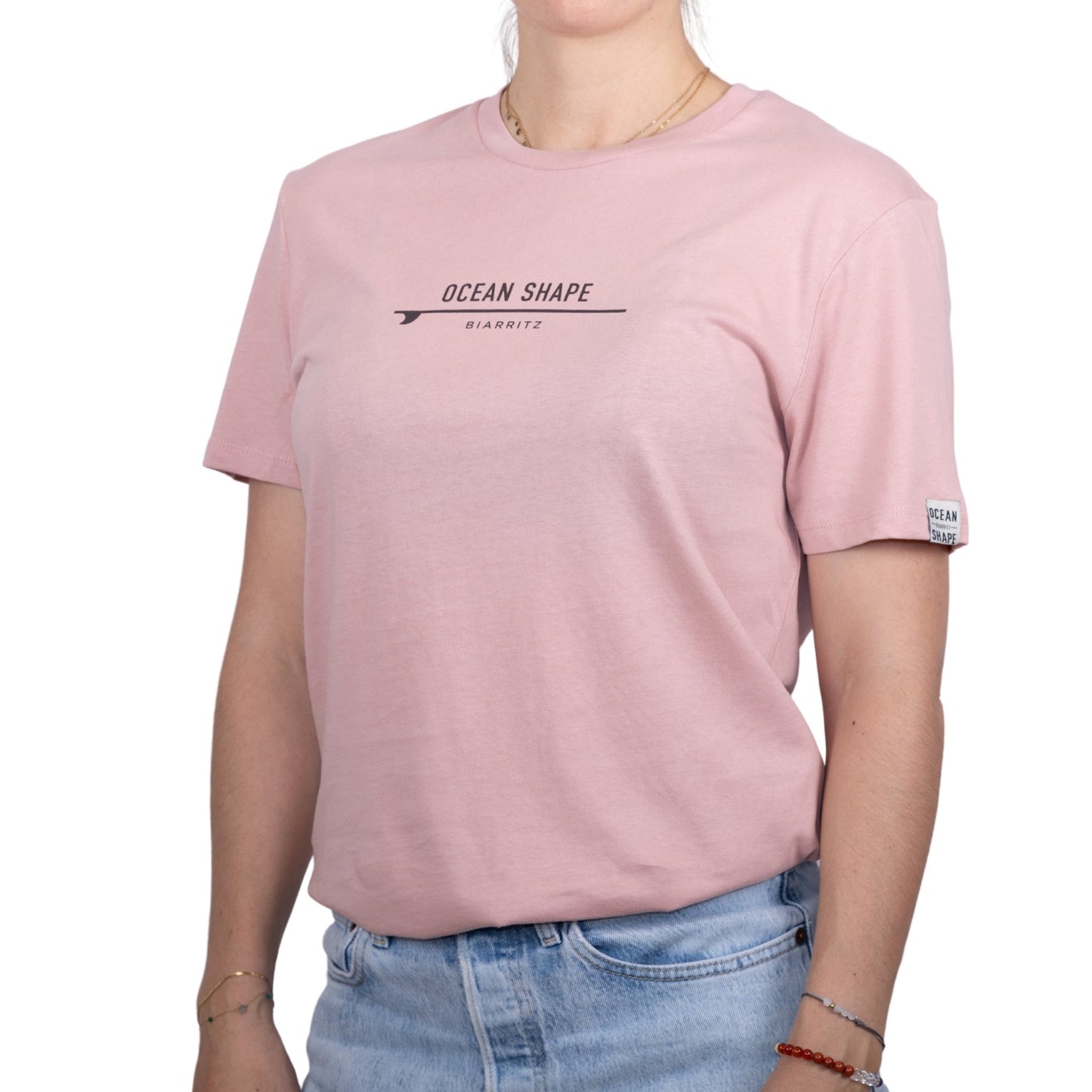 Tee-shirt unisexe rose pale en coton biologique avec petite signature "ocean shape Biarritz" sur surf marron foncé sur la poitrine, vu de face sur mannequin femme