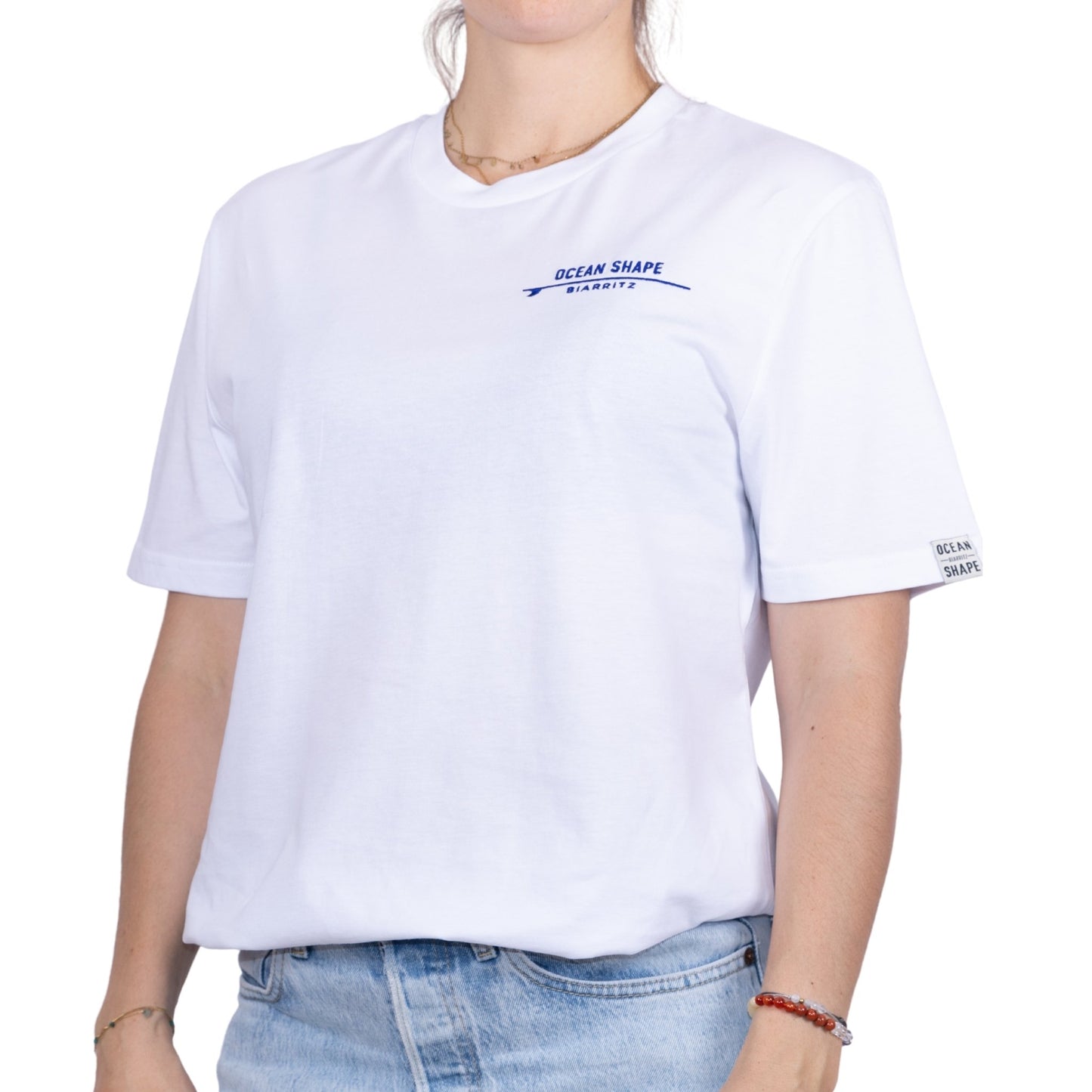 T-shirt unisexe blanc en coton biologique avec logo signature « ocean shape Biarritz » sur planche de surf minimaliste bleu marine, vue de face sur mannequin femme