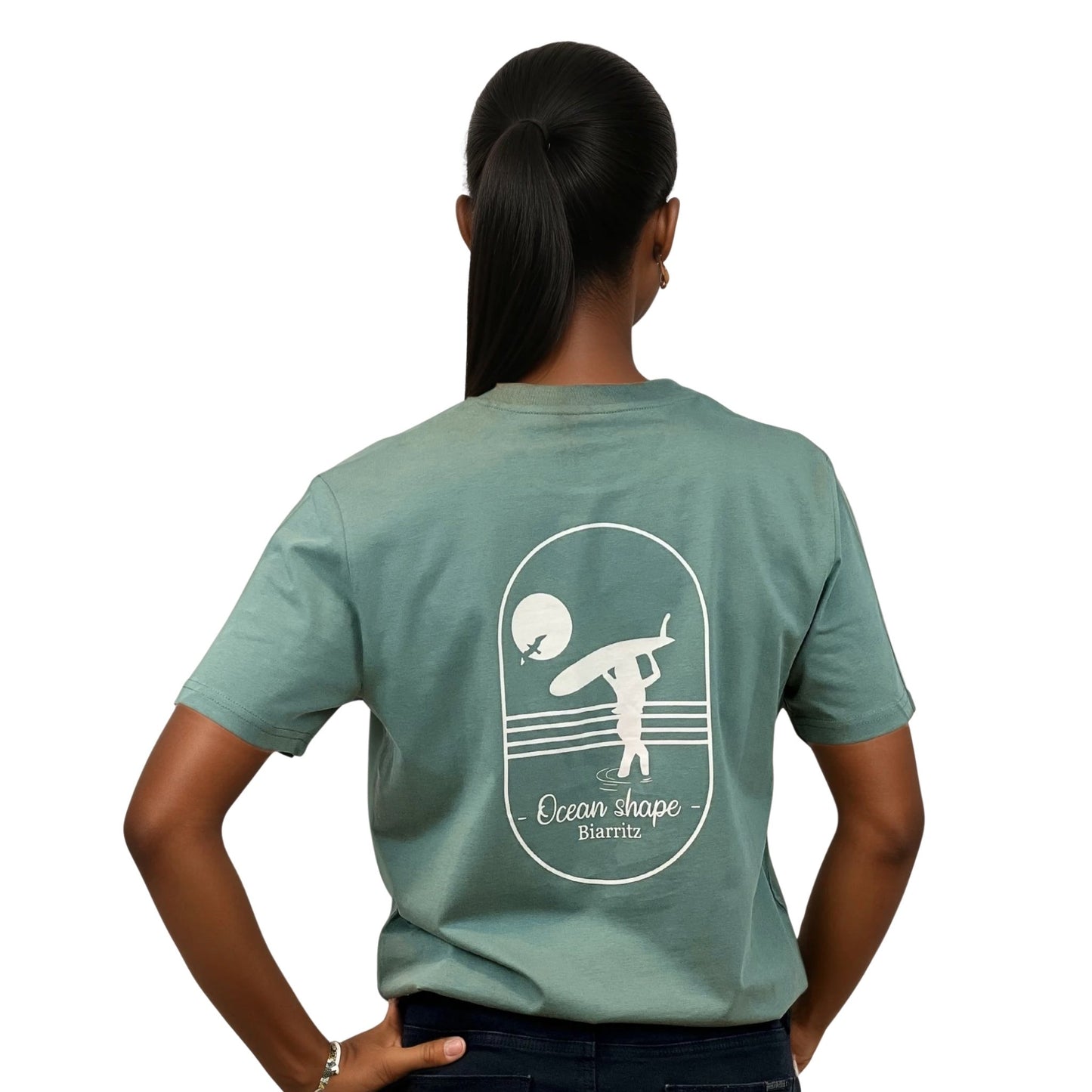 Tee-shirt unisexe vert sauge en coton biologique avec grand dessin de surfeuse qui marche + écriture "Ocean Shape Biarritz" écru dans le dos, vu de dos sur mannequin femme.
