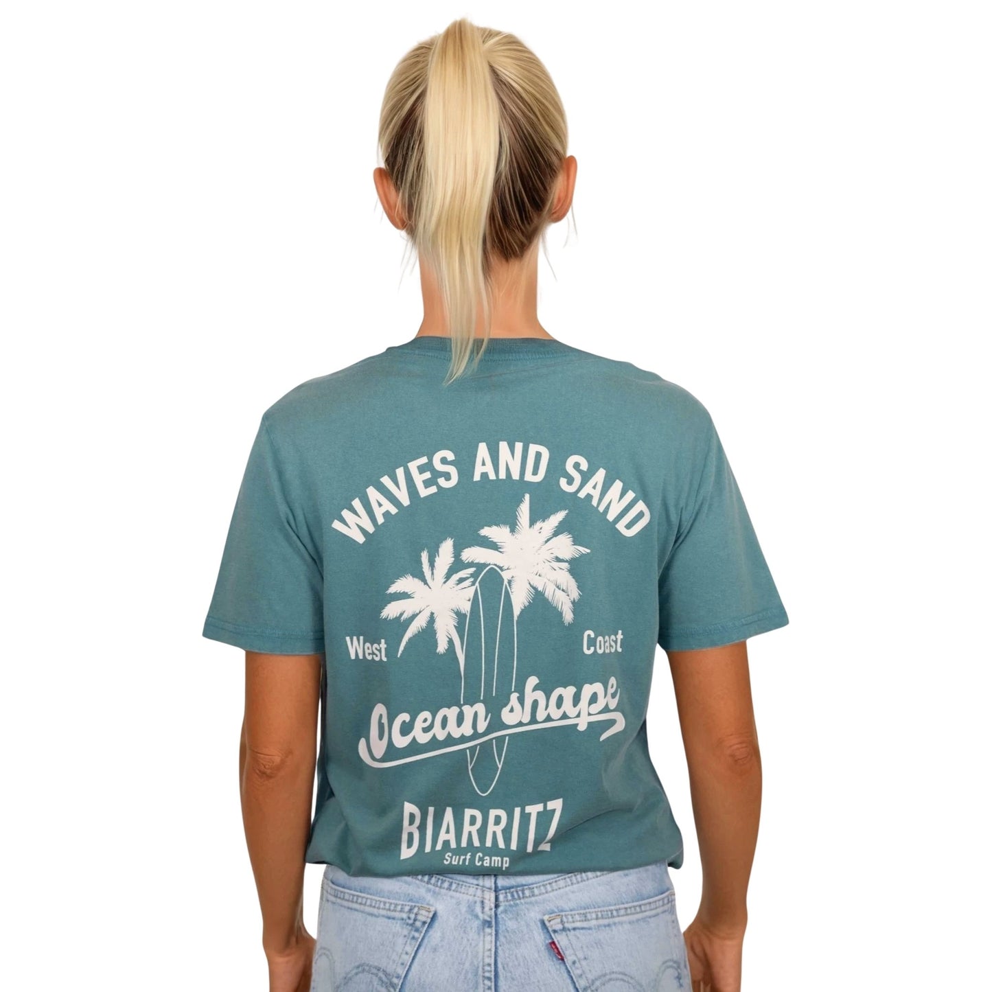 T-shirt unisexe en coton biologique vert sauge délavé avec grand dessin de palmiers, planche de surf et écriture « Waves and sand » et « Ocean Shape Biarritz » écru dans le dos, vu de dos sur mannequin femme.