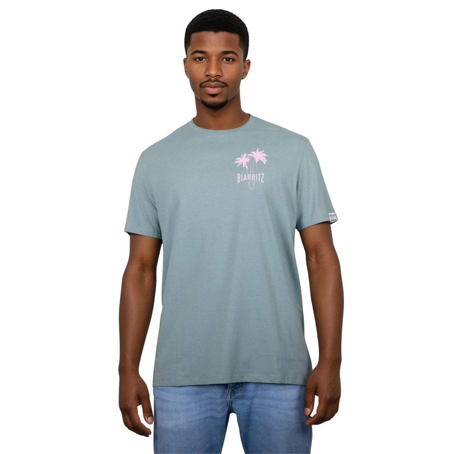 Tee-shirt unisexe vert sauge en coton biologique avec petit dessin de palmiers, planche de surf et écriture "Biarritz" rose clair sur la poitrine, vu de face sur mannequin homme.