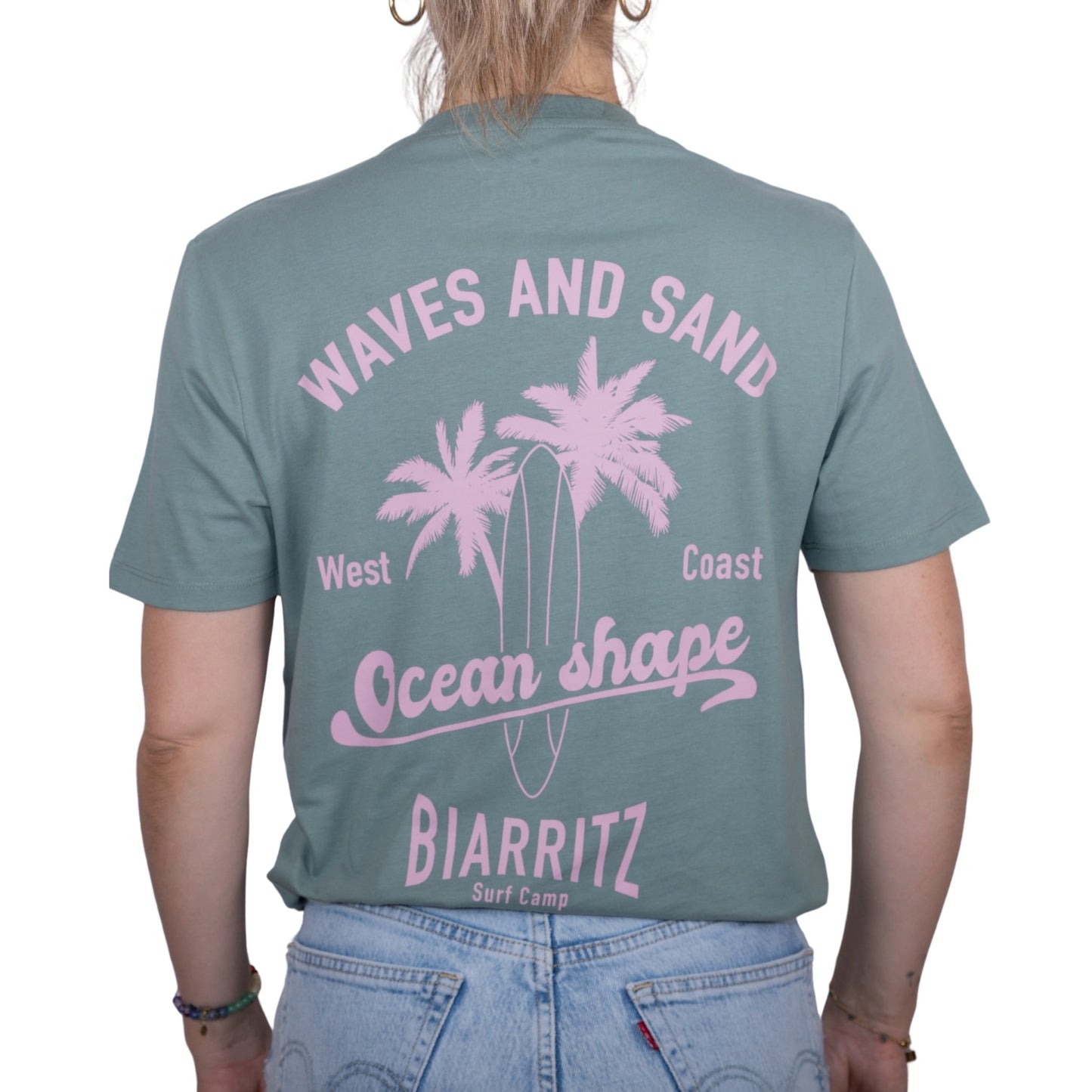Tee-shirt unisexe vert sauge en coton biologique avec grand dessin de palmiers, planche de surf et écriture "Waves and Sand" et "Ocean Shape Biarritz" rose clair dans le dos, vu de dos sur mannequin femme