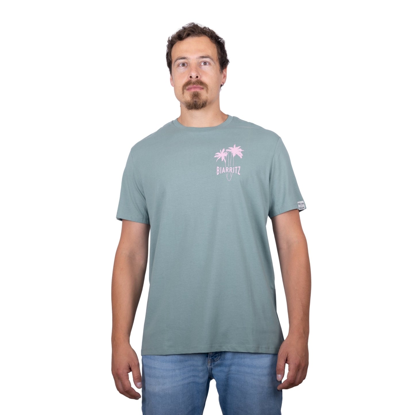 Tee-shirt unisexe vert sauge en coton biologique avec petit dessin de palmiers, planche de surf et écriture "Biarritz" rose clair sur la poitrine, vu de face sur mannequin homme