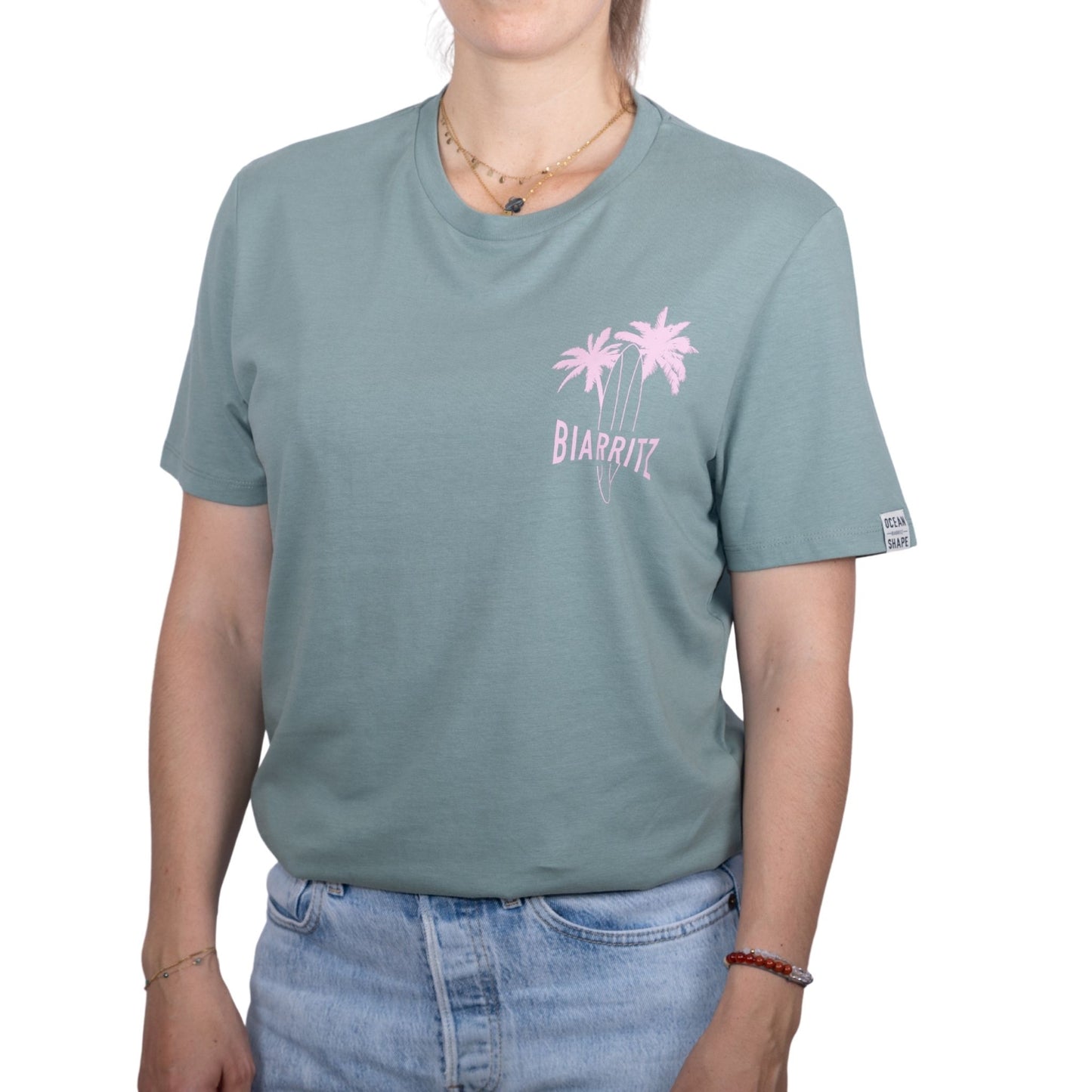 Tee-shirt unisexe vert sauge en coton biologique avec petit dessin de palmiers, planche de surf et écriture "Biarritz" rose clair sur la poitrine, vu de face sur mannequin femme