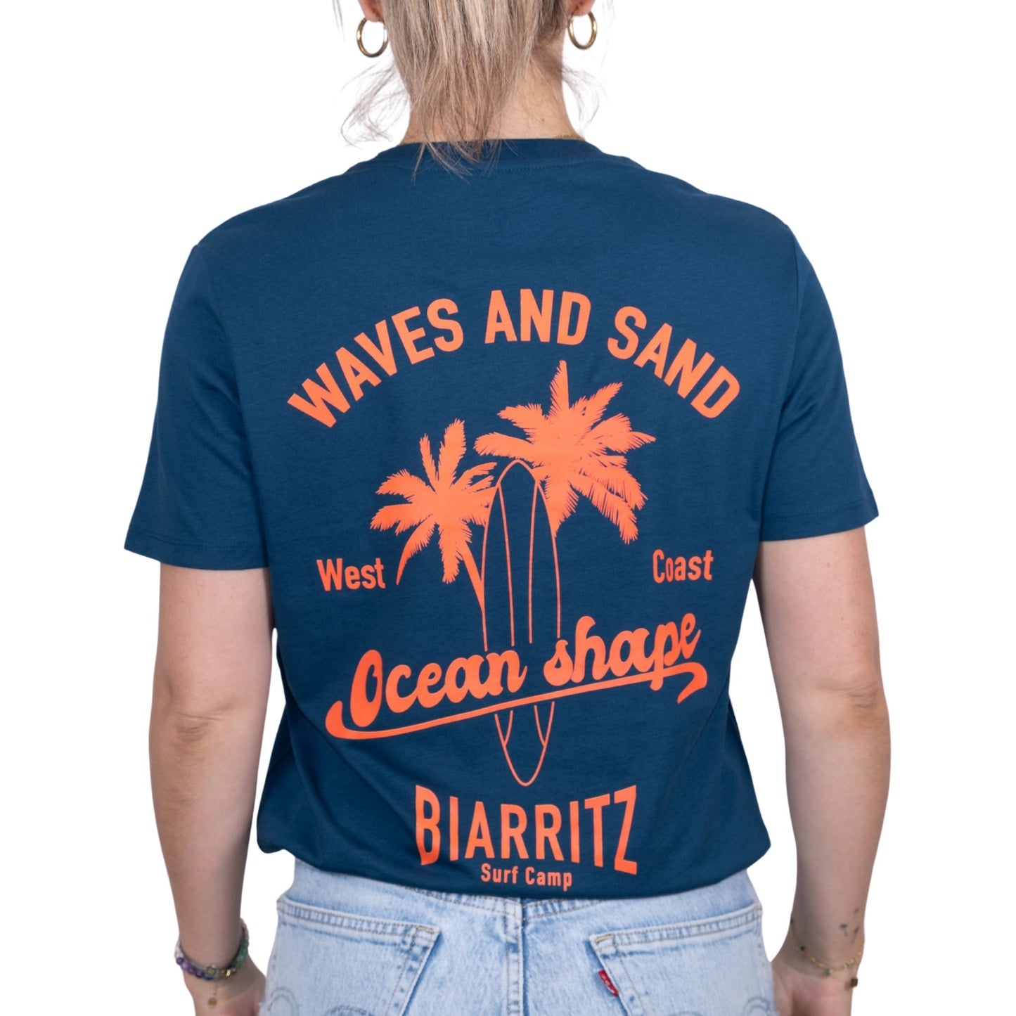 T-shirt unisexe en coton biologique bleu pétrole avec gros dessin palmiers, planche de surf et écriture « ocean shape Biarritz » et « waves and sun » en orange fluo dans le dos, vu de dos sur mannequin femme