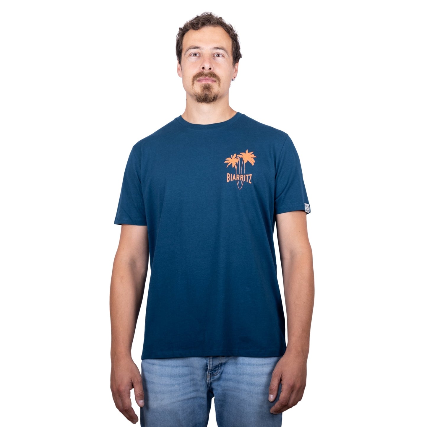 T-shirt unisexe en coton biologique bleu pétrole avec petit dessin palmiers, planche de surf et écriture « Biarritz » en orange fluo sur la poitrine, vu de face sur mannequin homme
