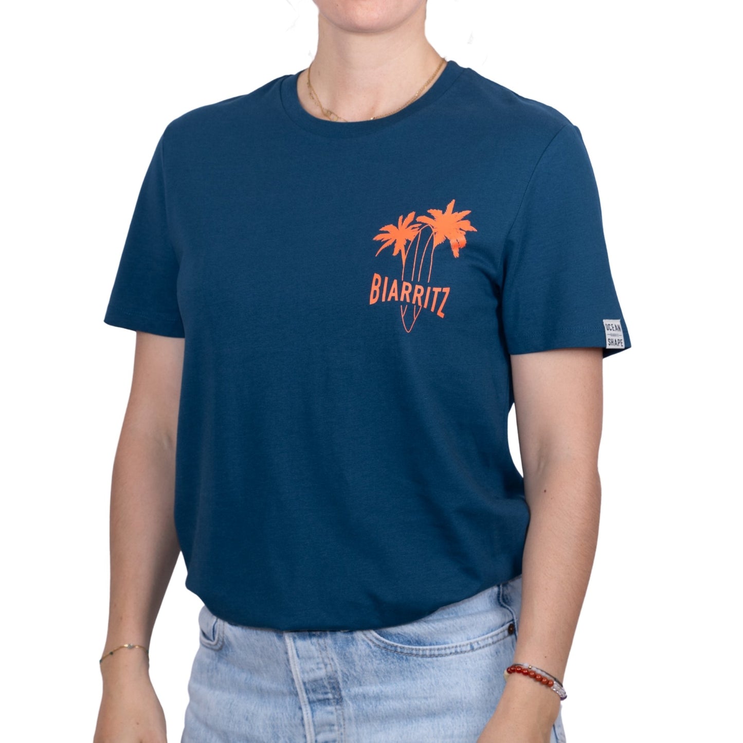 T-shirt unisexe en coton biologique bleu pétrole avec petit dessin palmiers, planche de surf et écriture « Biarritz » en orange fluo sur la poitrine, vu de face sur mannequin femme
