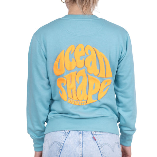 Sweat unisexe sans capuche Teal Monstera avec grand logo bubble "ocean shape biarritz" orange fluo dans le dos, vu de dos sur mannequin femme.