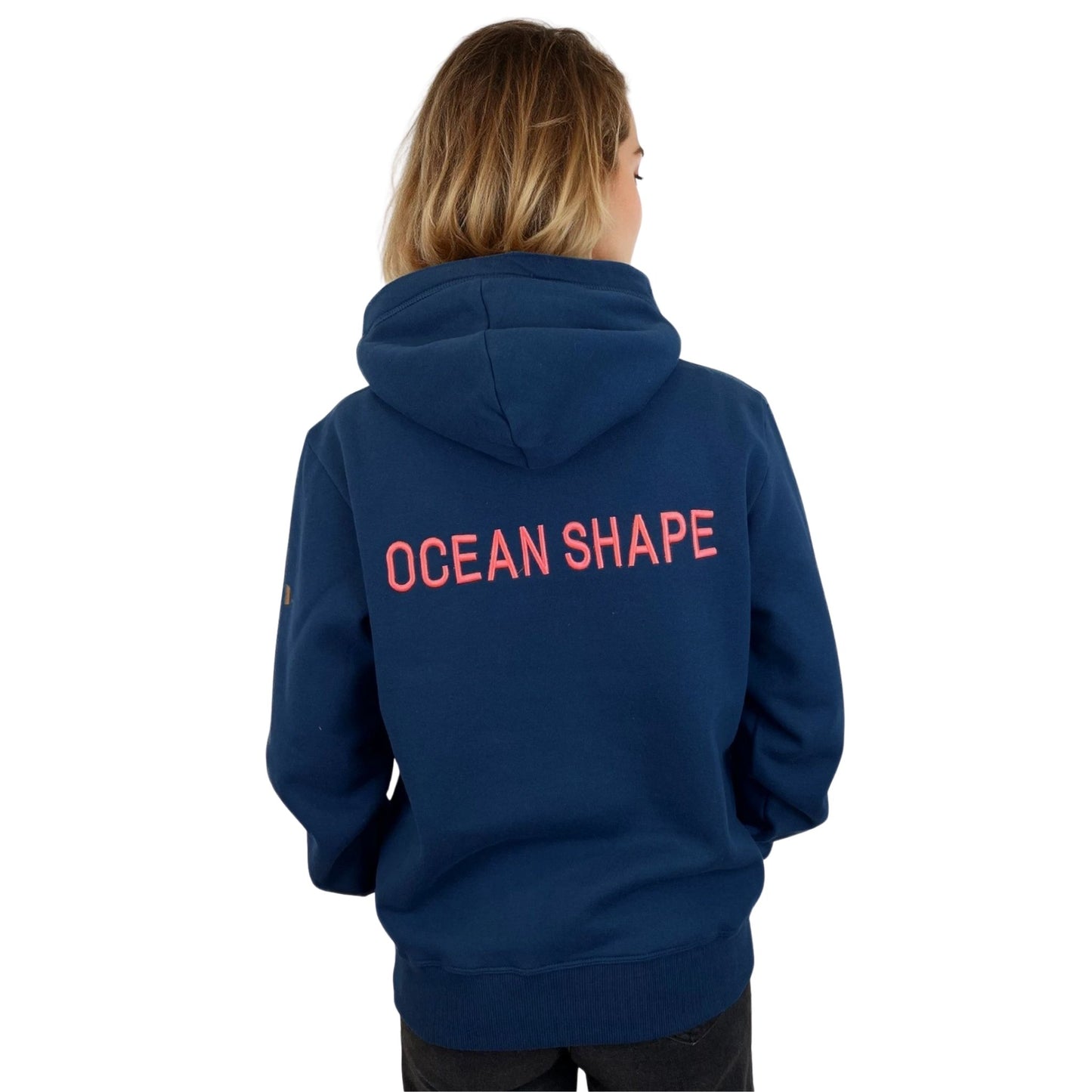 Sweat à capuche unisexe OceanShape couleur bleu paon, porté par une femme de dos.
Inscription "OceanShape" en majuscule, de couleur rose et brodé au milieu du dos.
Style casual et surf, inspiré de la côte basque.