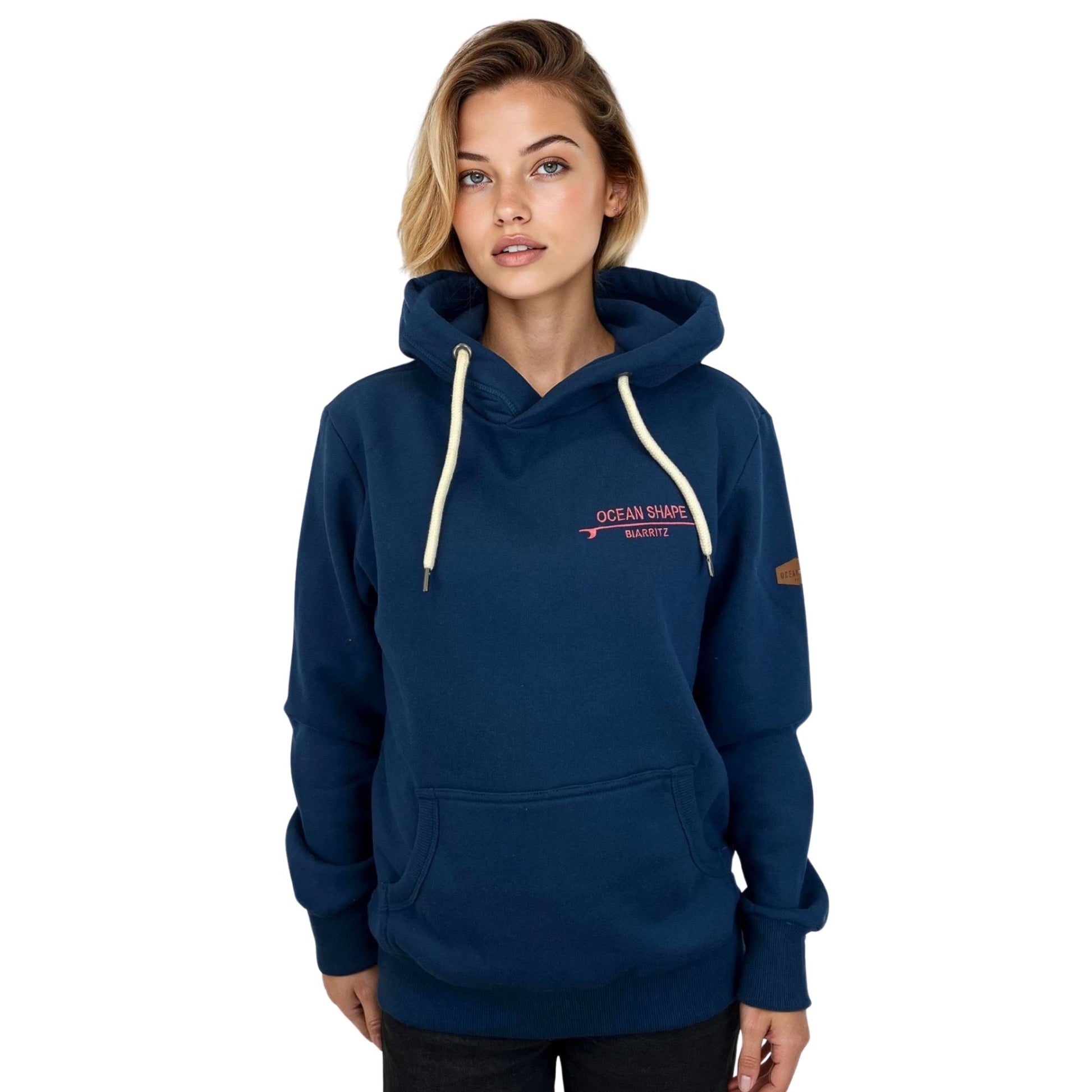 Sweat à capuche unisexe OceanShape couleur bleu paon, porté par une femme de face.
Inscription "OceanShape" avec la mention "Biarritz" brodé en rose, et en majuscule.
Style casual et surf, inspiré de la côte basque.