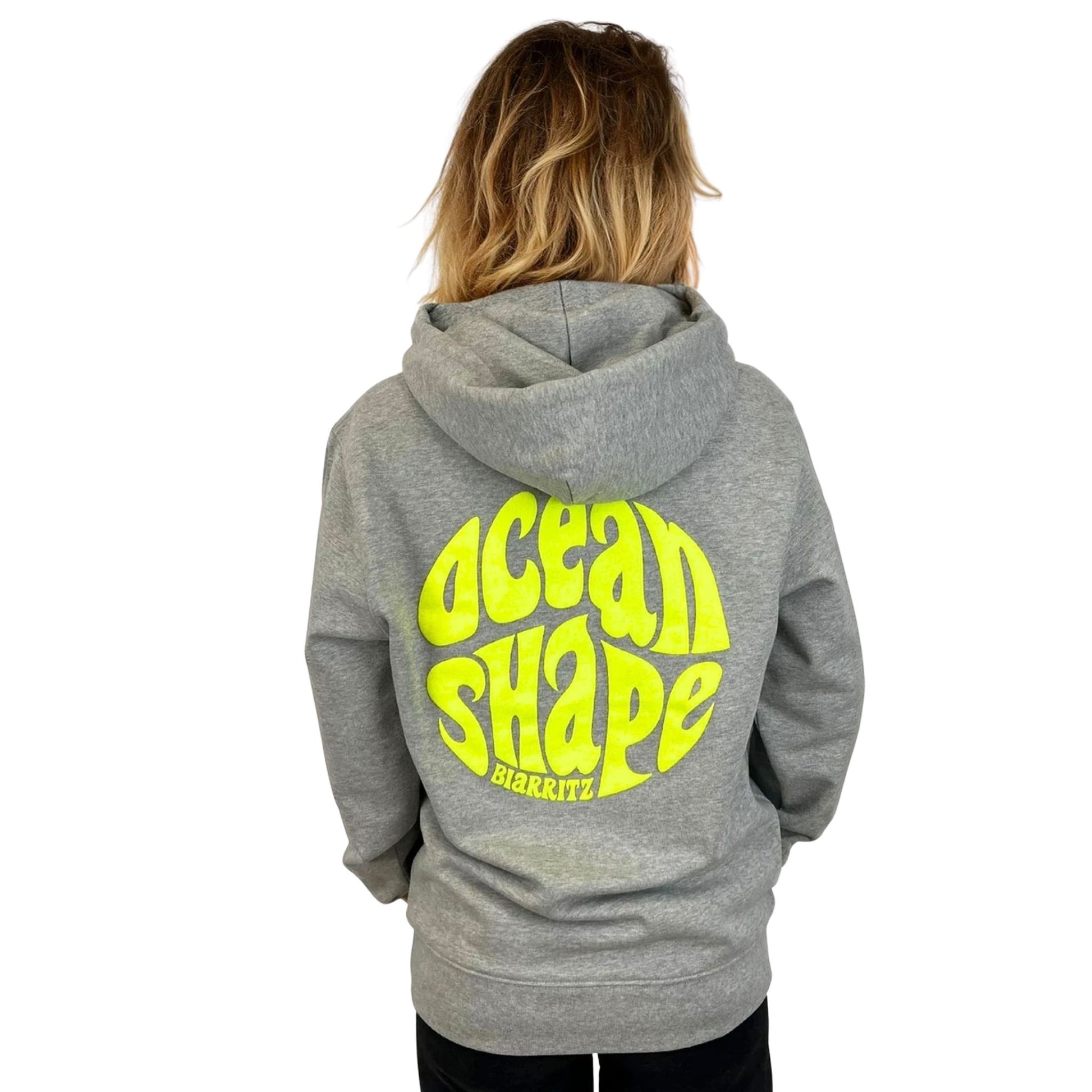 Sweat à capuche unisexe gris.
Gros logo bubble "Ocean Shape Biarritz" en encre gonflante jaune fluo dans le dos.
Vu de dos sur mannequin femme.