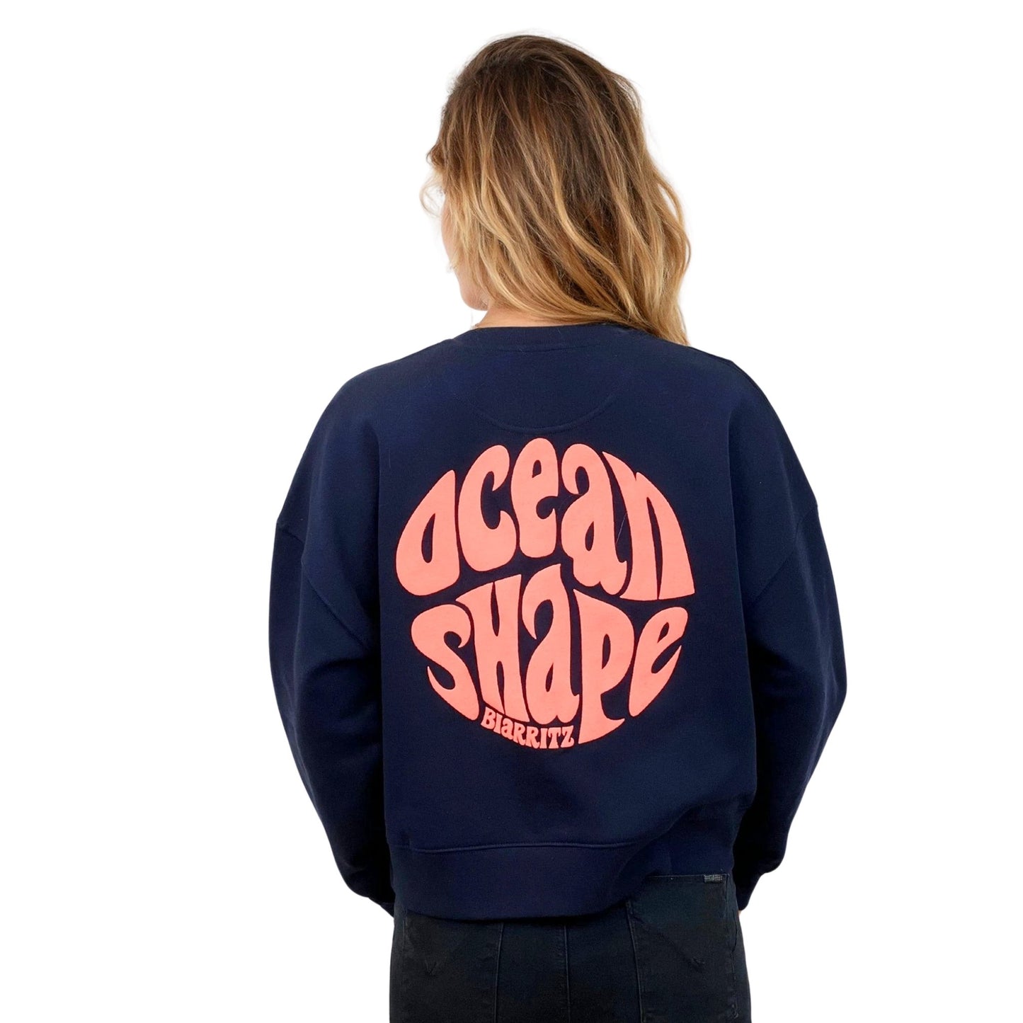 Sweat femme court et oversize, col rond, épaules tombantes, 100 % coton biologique, bleu marine.
Gros logo bubble "Ocean shape Biarritz" corail fluo.
Vu de dos sur mannequin femme.