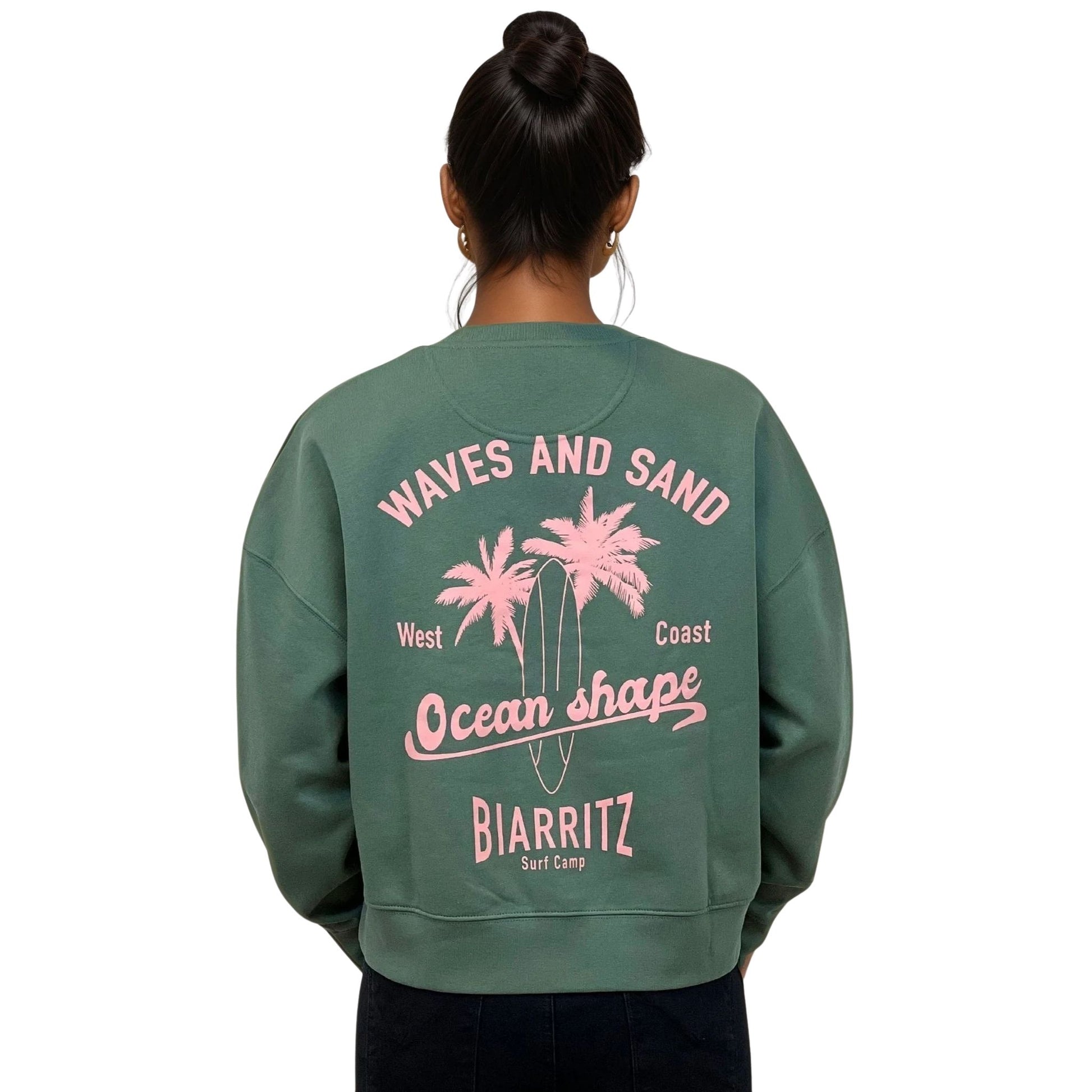 Sweat femme court et oversize, col rond, épaules tombantes, 100 % coton biologique, couleur vert sauge.
Grand dessin rose pâle de planche de surf et palmiers + écriture "Waves and sand, Ocean Shape Biarritz" dans le dos.
Vu de dos sur mannequin femme.