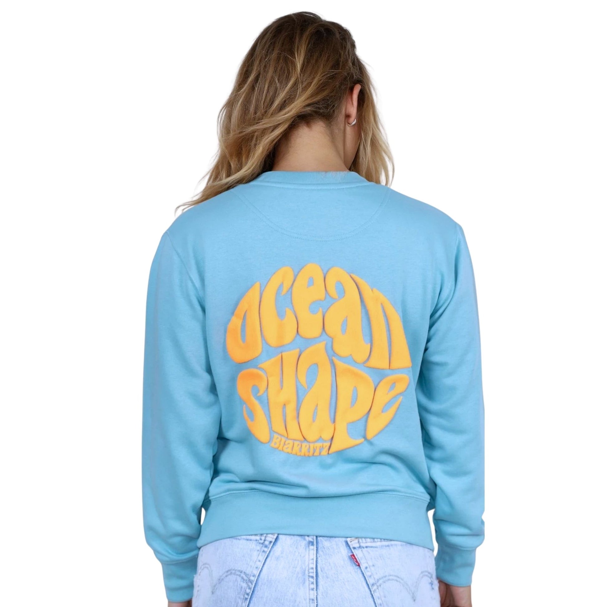 Sweat unisexe sans capuche Teal Monstera avec grand logo bubble "ocean shape biarritz" orange fluo dans le dos, vu de dos sur mannequin femme.