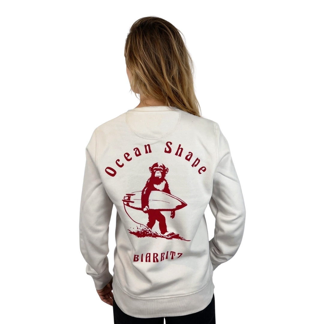 Sweat sans capuche unisexe OceanShape crème en coton biologique avec gros dessin de singe surfeur et écriture « ocean shape Biarritz » rouge dans le dos, vu de dos sur mannequin femme.