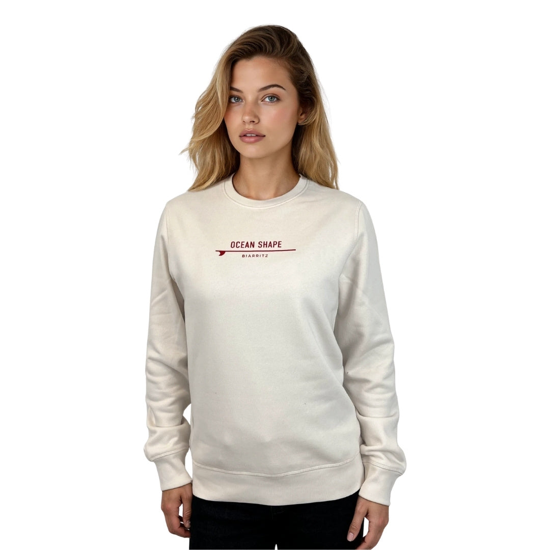 Sweat à col rond unisexe OceanShape de couleur crème en coton biologique avec petite signature "ocean shape Biarritz" sur surf rouge sur la poitrine, vu de face sur mannequin femme.
