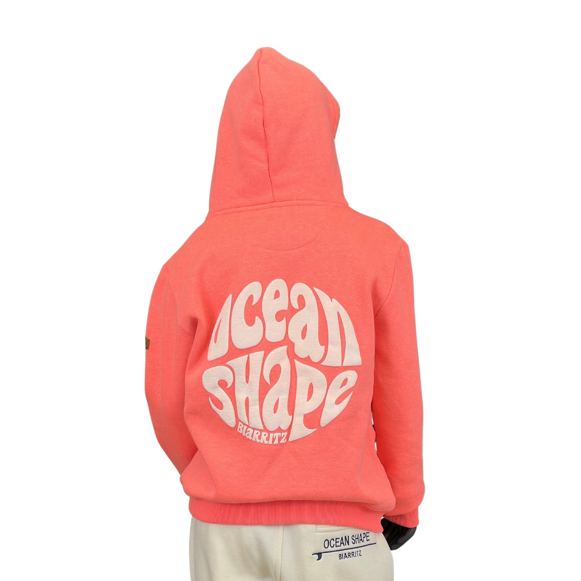 sweat à capuche unisexe enfant, en coton biologique ocean shape de couleur corail sur un mannequin enfant de dos. 
Dos : grand logo bubble ocean shape blanc en relief avec écrit biarritz.