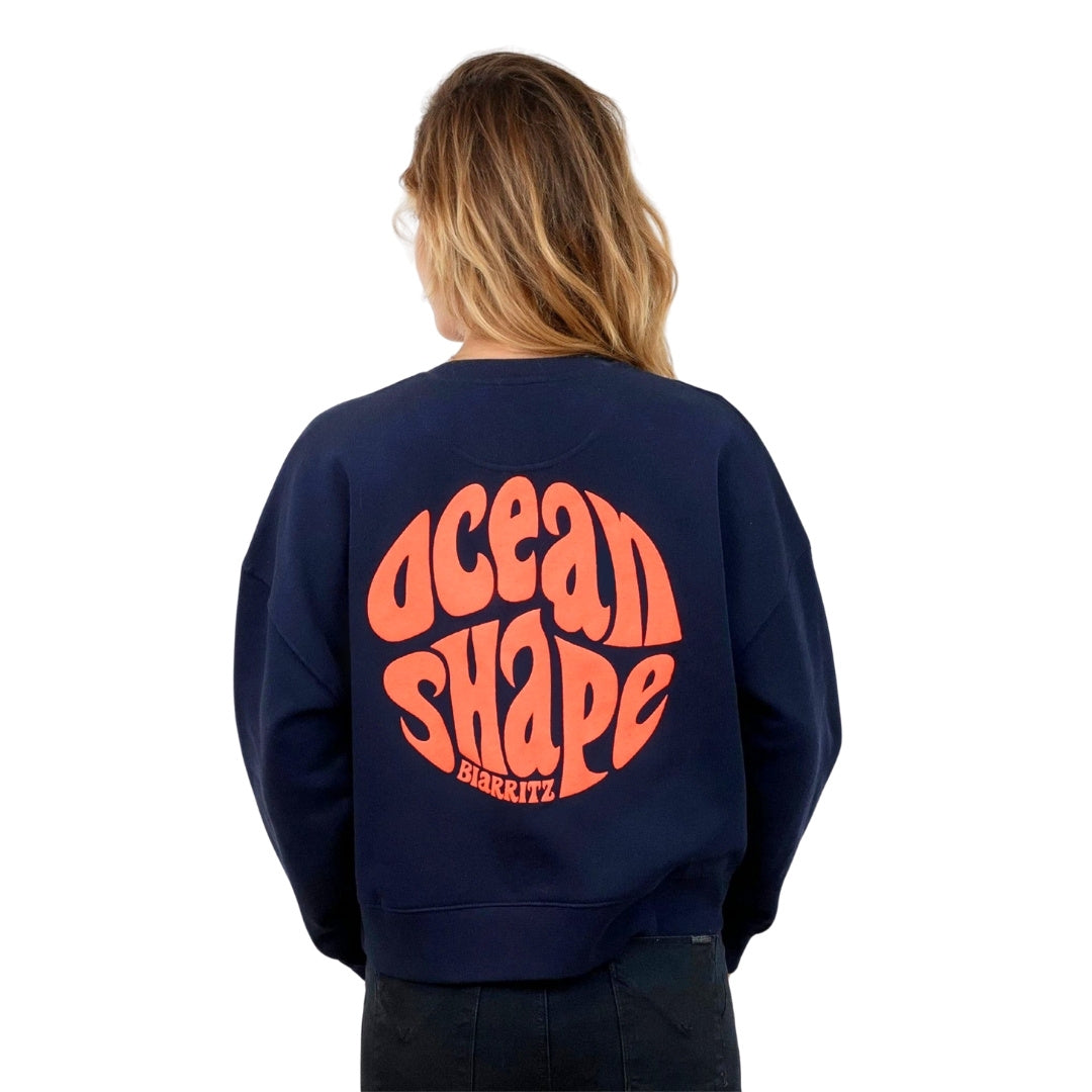 Sweat femme court et oversize, col rond, épaules tombantes, 100 % coton biologique, bleu marine.
Gros logo bubble "Ocean shape Biarritz" corail fluo.
Vu de dos sur mannequin femme.