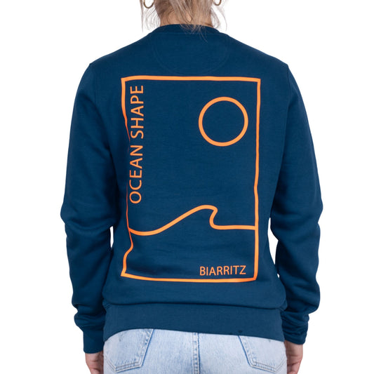 sweat unisexe bleu petrole en coton biologique avec grand encadré vague / soleil minimaliste + écriture "ocean shape Biarritz" orange fluo dans le dos, vu de dos sur mannequin femme.