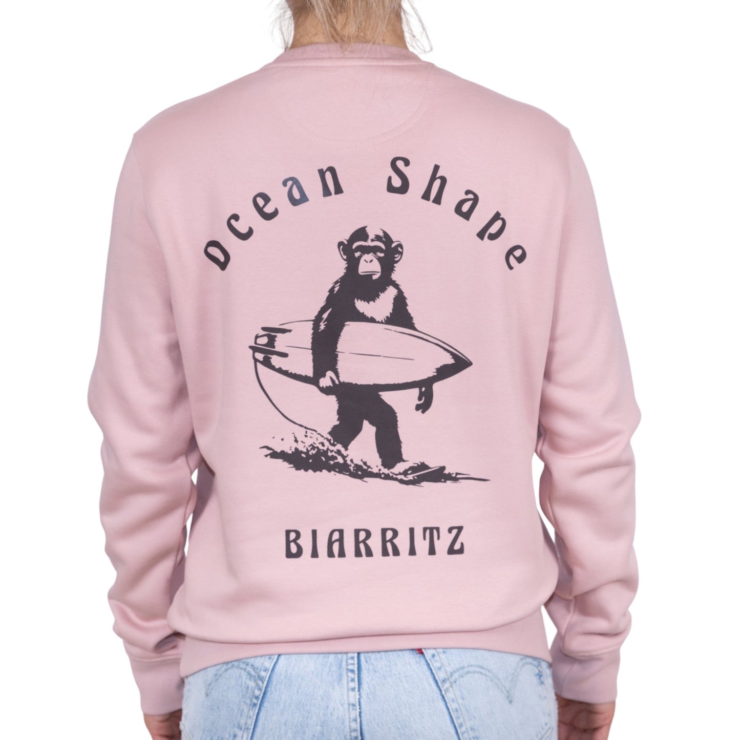 Sweat unisexe rose pale en coton biologique avec gros dessin de singe surfeur et écriture « ocean shape Biarritz » marron foncé dans le dos, vu de dos sur mannequin femme.