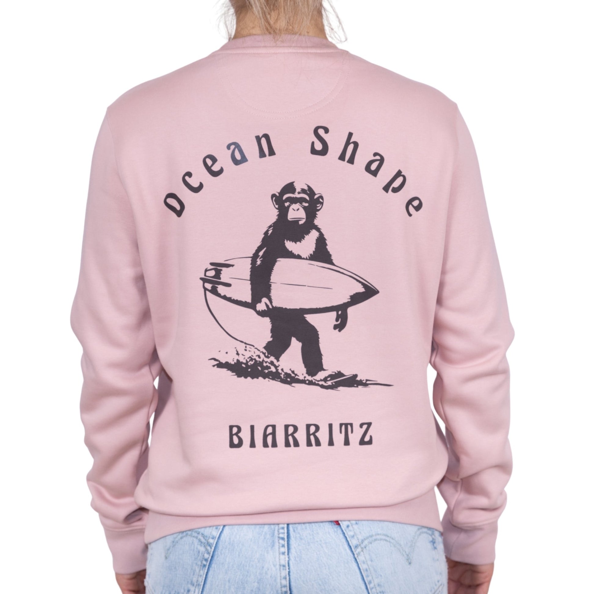 Sweat unisexe rose pale en coton biologique avec gros dessin de singe surfeur et écriture « ocean shape Biarritz » marron foncé dans le dos, vu de dos sur mannequin femme.