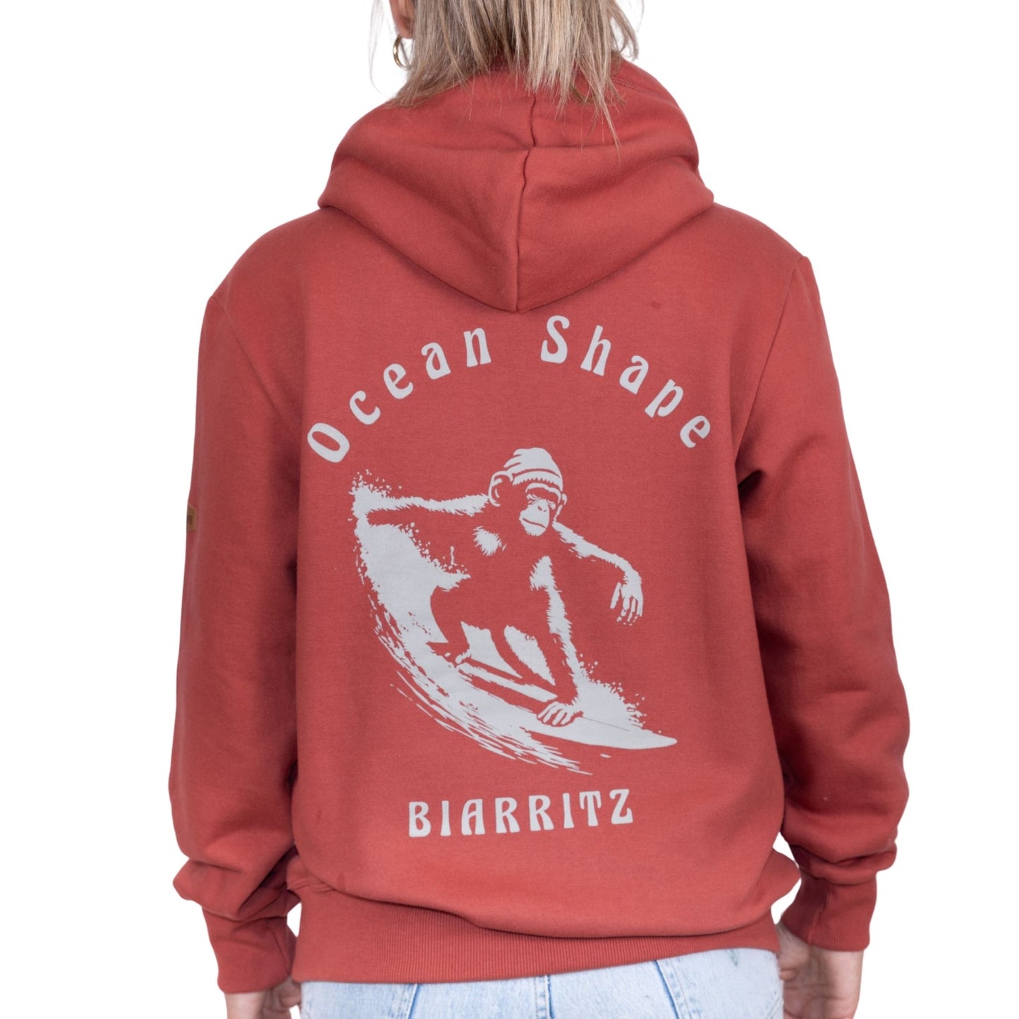 Sweat unisexe à capuche terracotta avec grand dessin de singe qui surf + écriture "Ocean Shape Biarritz" beige dans le dos, vu de dos sur mannequin femme