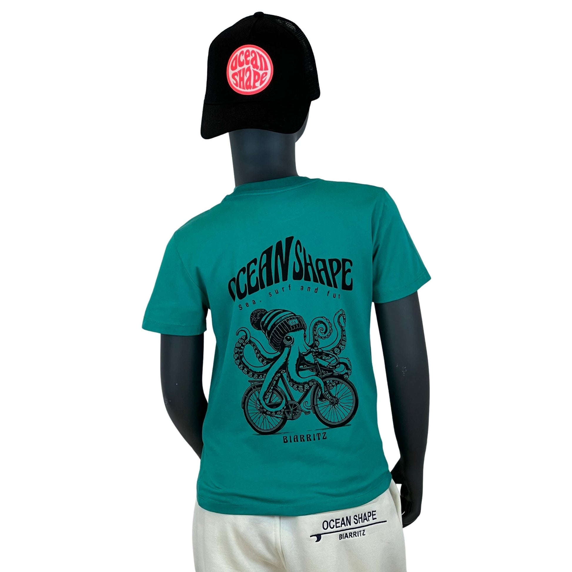 t-shirt enfant à manches courtes unisexe ocean shape bleu vert, 100% coton biologique. 
Gros dessin noir d'une pieuvre avec un bonnet qui fait du vélo avec écriture "ocean shape biarritz et sea, surf and fun" dans le dos.
Vu de dos sur mannequin enfant