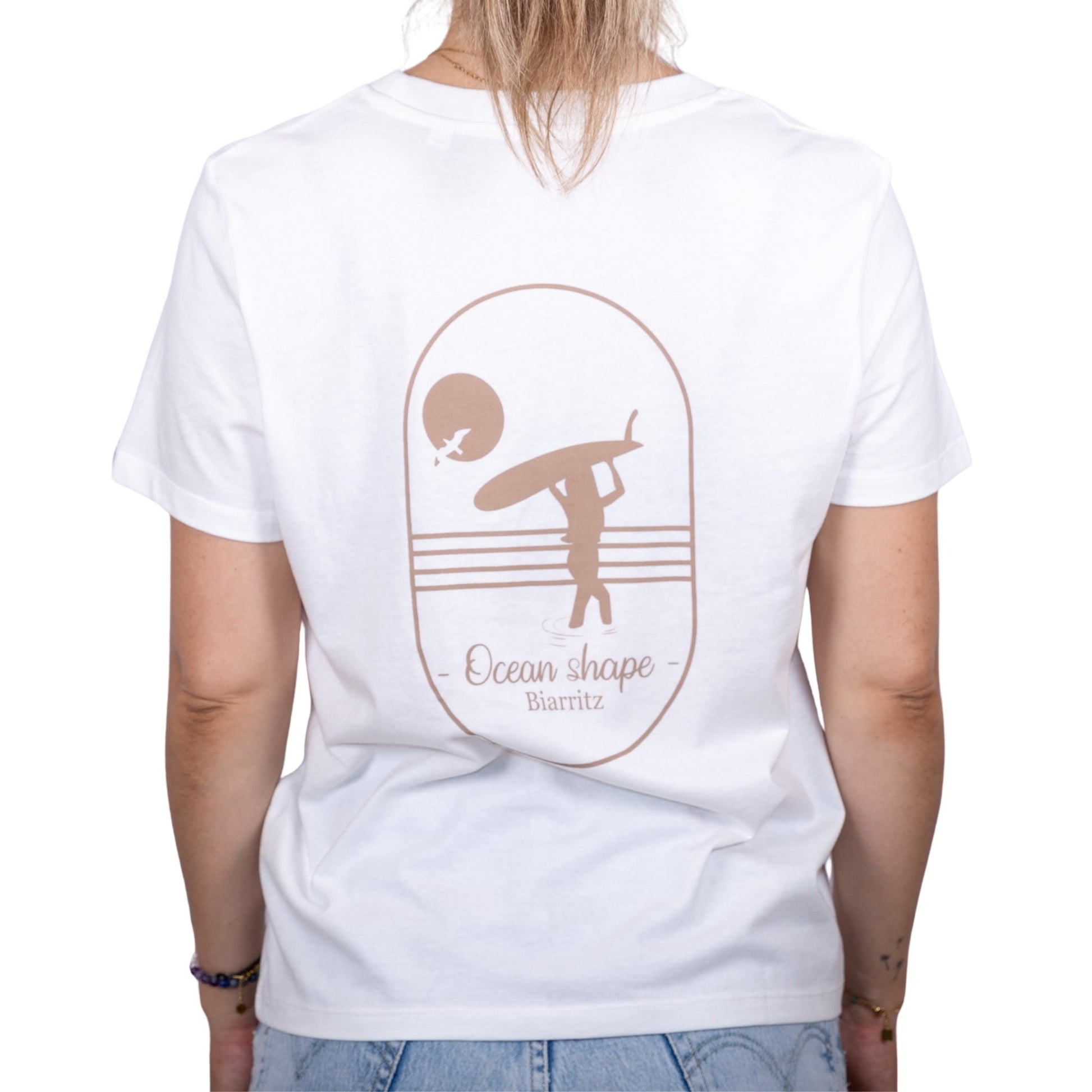 Tee-shirt femme en coton biologique blanc avec gros dessin de surfeuse qui marche de dos et écriture "ocean shape Biarritz" beige dans le dos , vu de dos sur mannequin femme