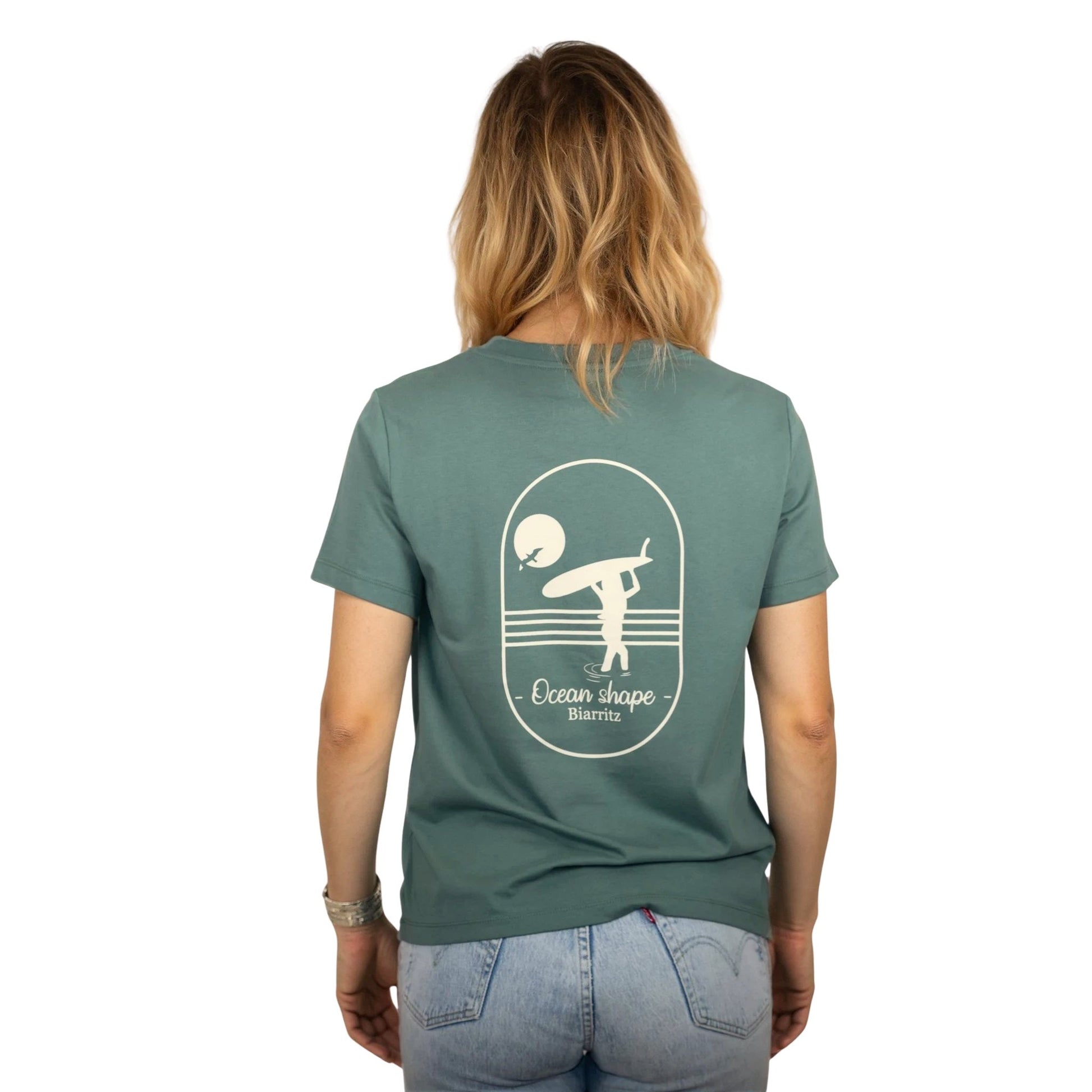 Tee-shirt femme en coton biologique vert sauge avec gros dessin surfeuse qui marche de dos et écriture ocean shape Biarritz écru dans le dos , vu de dos sur mannequin femme 