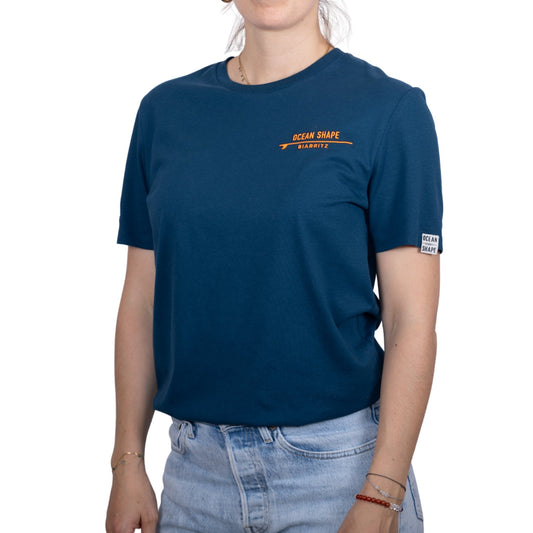 T-shirt unisexe bleu pétrole en coton biologique avec logo signature « ocean shape Biarritz » sur planche de surf minimaliste orange fluo, vue de face sur mannequin femme