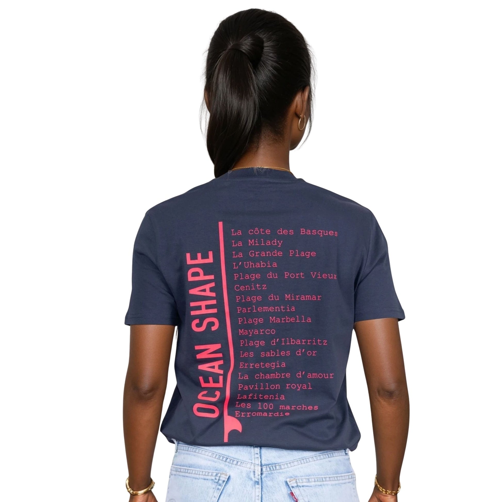 Tee-shirt unisexe gris foncé en coton biologique avec longue liste de plages et grande écriture "ocean shape" rose fluo dans le milieu du dos, vu de dos sur mannequin femme.