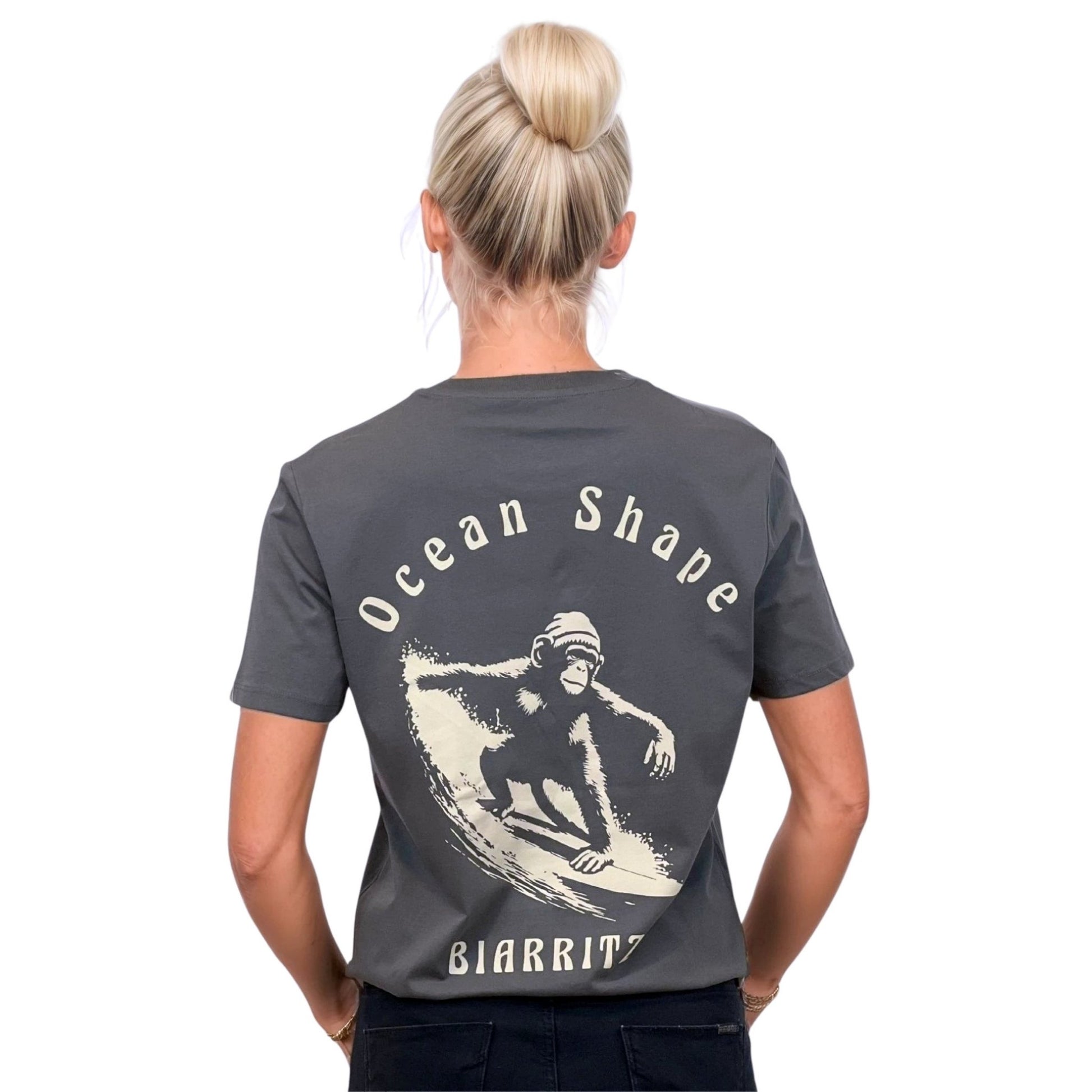 Tee-shirt unisexe gris foncé en coton biologique avec grand dessin de singe qui surfe et écriture « ocean shape Biarritz » beige dans le dos, vu de dos sur mannequin femme.
