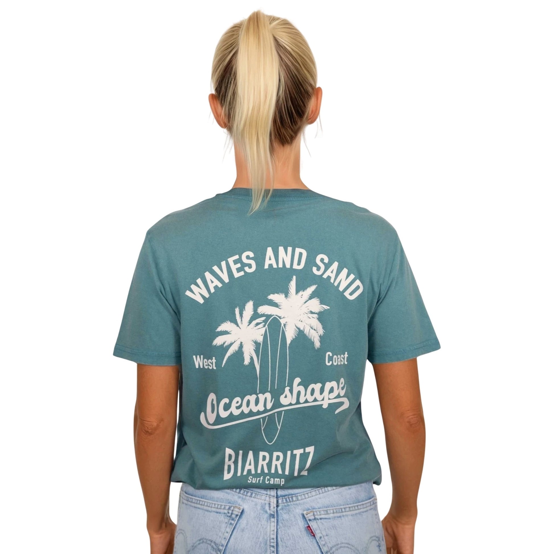 T-shirt unisexe en coton biologique vert sauge délavé avec grand dessin de palmiers, planche de surf et écriture « Waves and sand » et « Ocean Shape Biarritz » écru dans le dos, vu de dos sur mannequin femme.