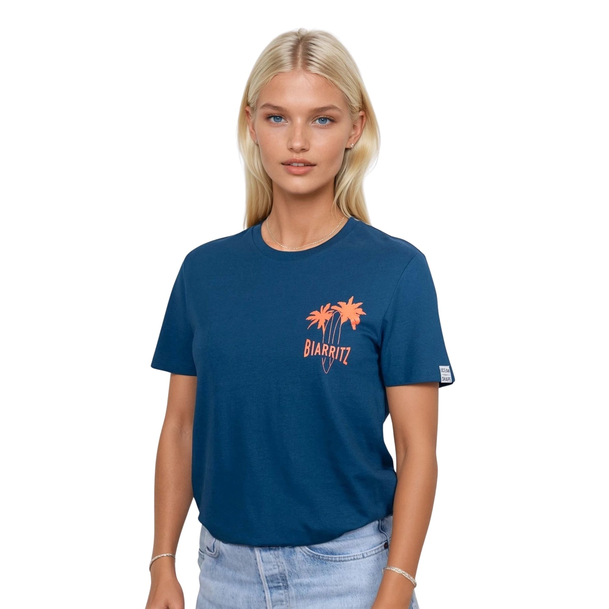 T-shirt unisexe en coton biologique bleu paon avec petit dessin palmiers, planche de surf et écriture « Biarritz » orange fluo sur la poitrine gauche, vu de face sur mannequin femme.