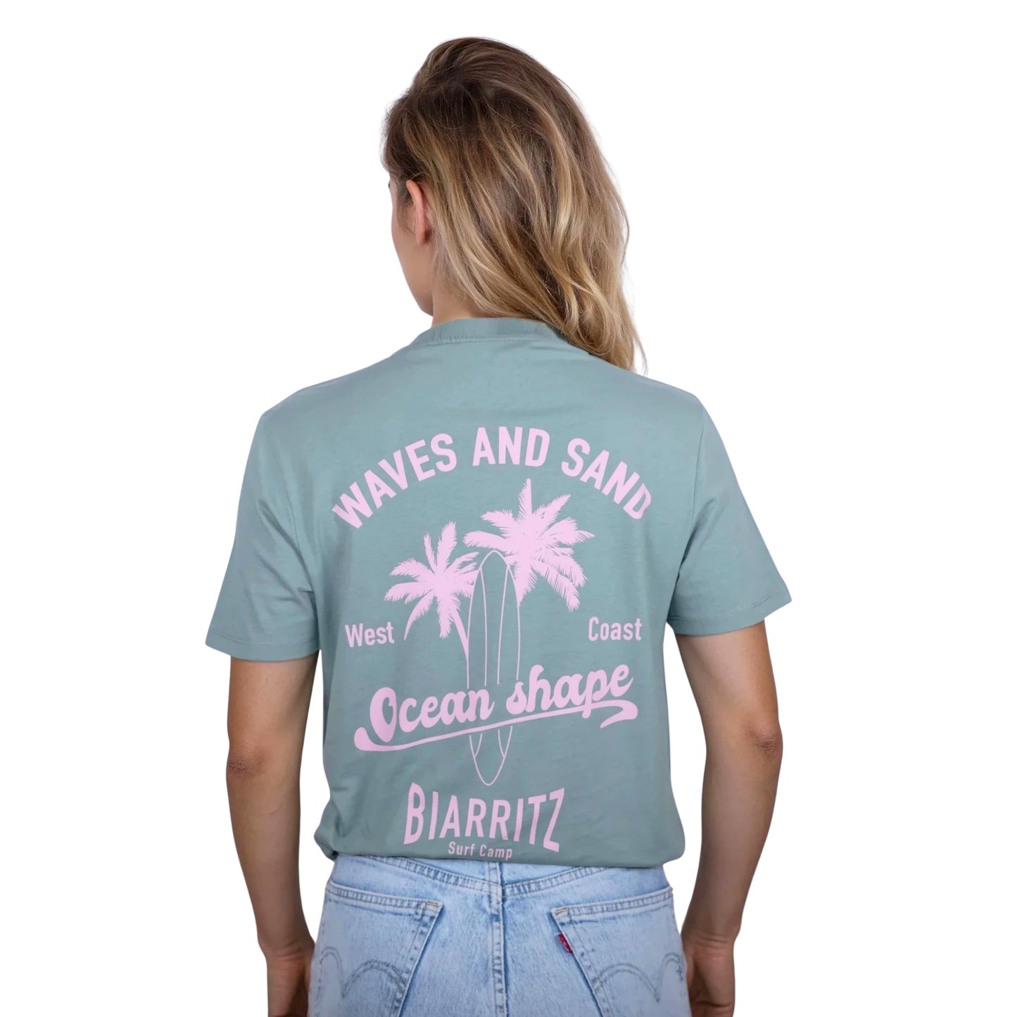 Tee-shirt unisexe vert sauge en coton biologique avec grand dessin de palmiers, planche de surf et écriture "Waves and Sand" et "Ocean Shape Biarritz" rose clair dans le dos, vu de dos sur mannequin femme.