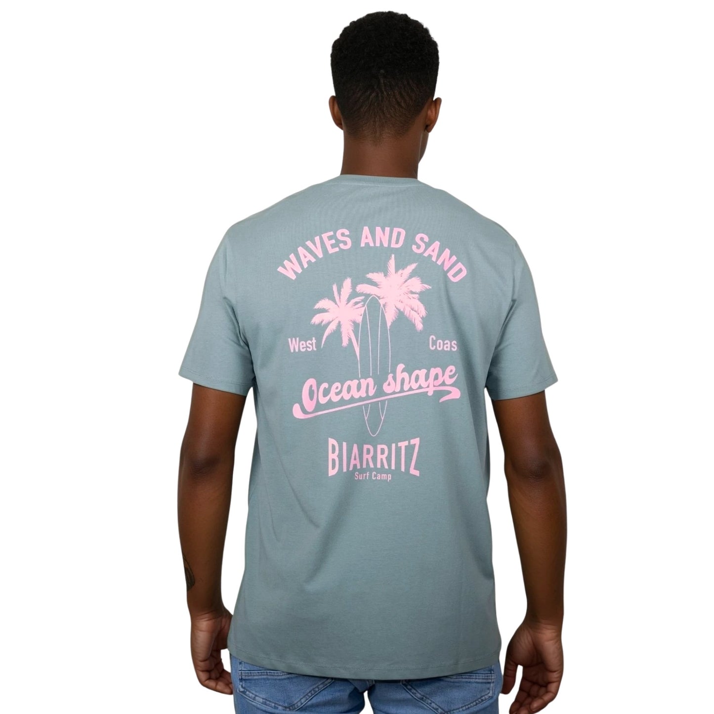 Tee-shirt unisexe vert sauge en coton biologique avec grand dessin de palmiers, planche de surf et écriture "Waves and Sand" et "Ocean Shape Biarritz" rose clair dans le dos, vu de dos sur mannequin homme.