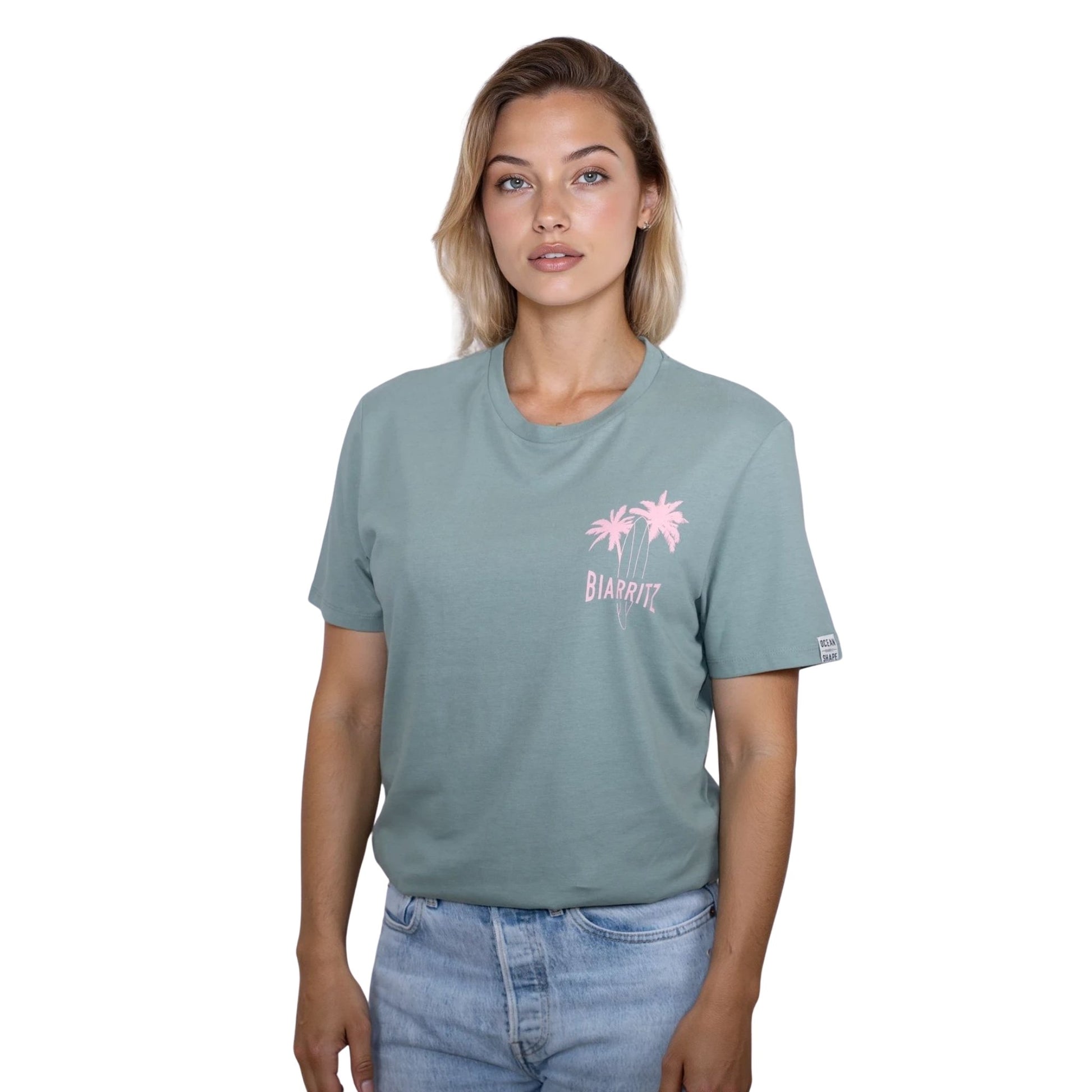 Tee-shirt unisexe vert sauge en coton biologique avec petit dessin de palmiers, planche de surf et écriture "Biarritz" rose clair sur la poitrine, vu de face sur mannequin femme.