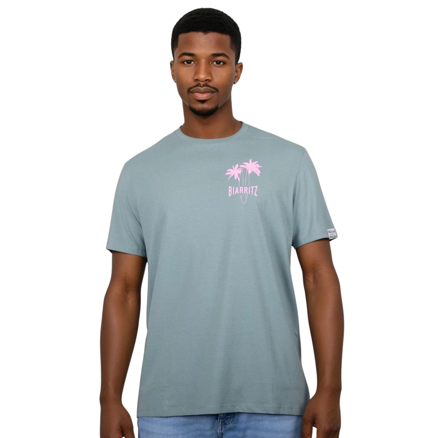 Tee-shirt unisexe vert sauge en coton biologique avec petit dessin de palmiers, planche de surf et écriture "Biarritz" rose clair sur la poitrine, vu de face sur mannequin homme.