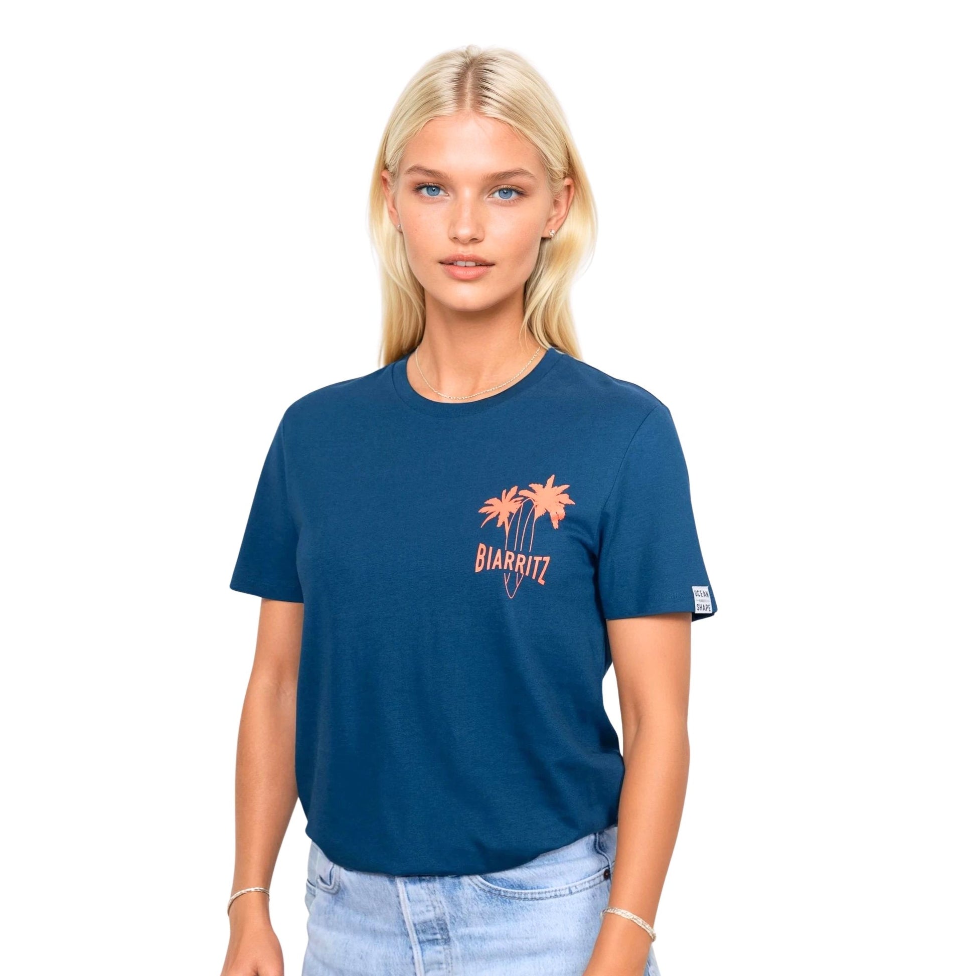 T-shirt unisexe en coton biologique bleu paon avec petit dessin palmiers, planche de surf et écriture « Biarritz » orange fluo sur la poitrine gauche, vu de face sur mannequin femme.