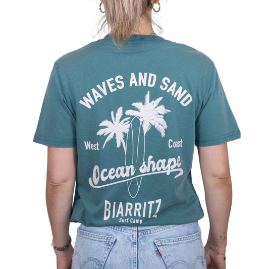 T-shirt unisexe en coton biologique vert sauge délavé avec grand dessin de palmiers, planche de surf et écriture « Waves and sand » et « Ocean Shape Biarritz » écru dans le dos, vu de dos sur mannequin femme