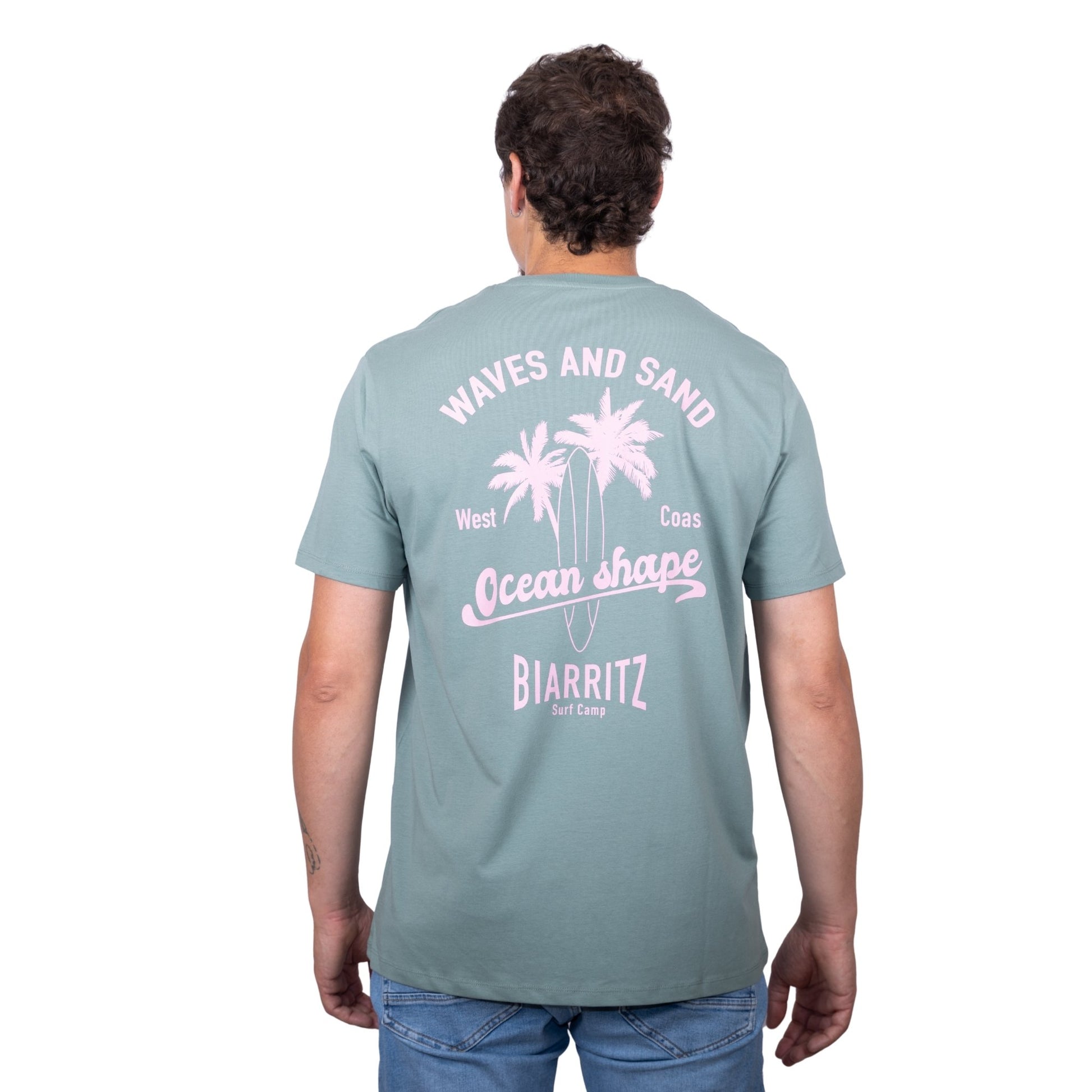 Tee-shirt unisexe vert sauge en coton biologique avec grand dessin de palmiers, planche de surf et écriture "Waves and Sand" et "Ocean Shape Biarritz" rose clair dans le dos, vu de dos sur mannequin homme