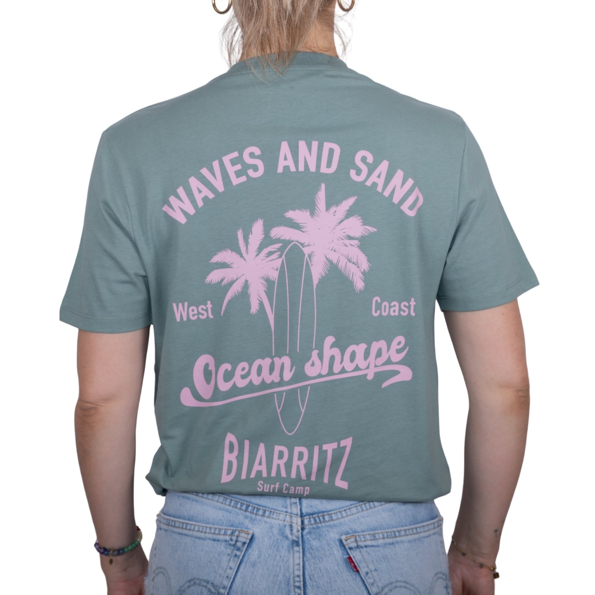 Tee-shirt unisexe vert sauge en coton biologique avec grand dessin de palmiers, planche de surf et écriture "Waves and Sand" et "Ocean Shape Biarritz" rose clair dans le dos, vu de dos sur mannequin femme